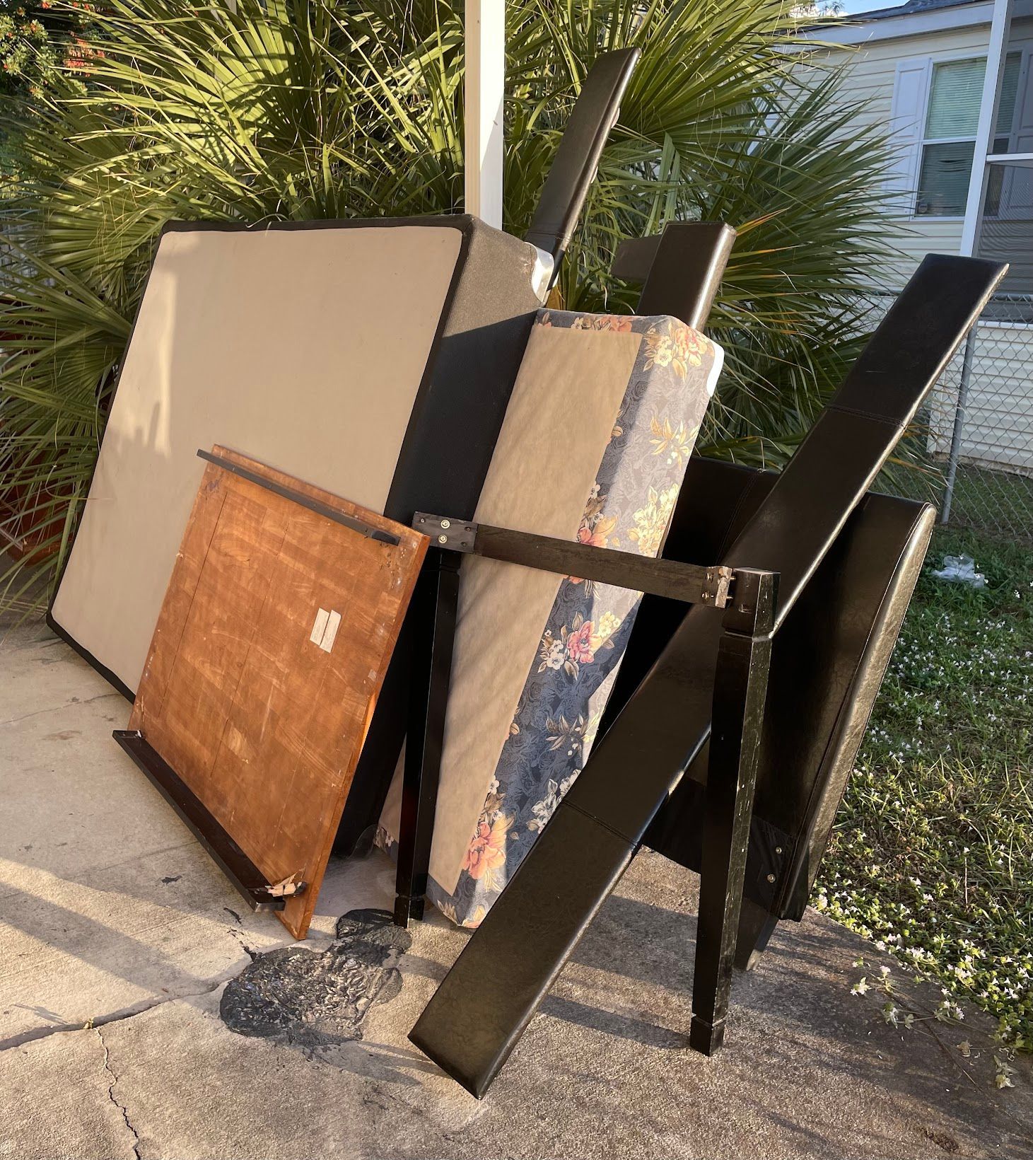 Pile of discarded furniture on a sidewalk, including a headboard, rolled rug, and chair.