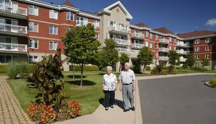 Elderly couple walking on a pathway in front of a retirement home on a sunny day.