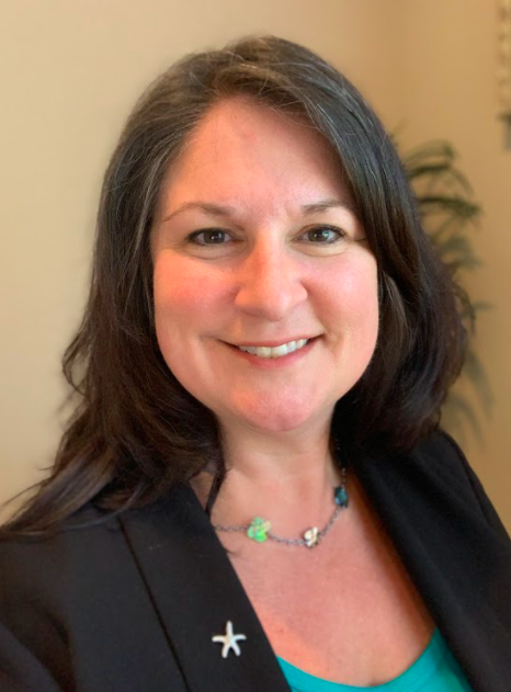 Smiling woman in a black blazer with a star pin, wearing a necklace, against a beige indoor background
