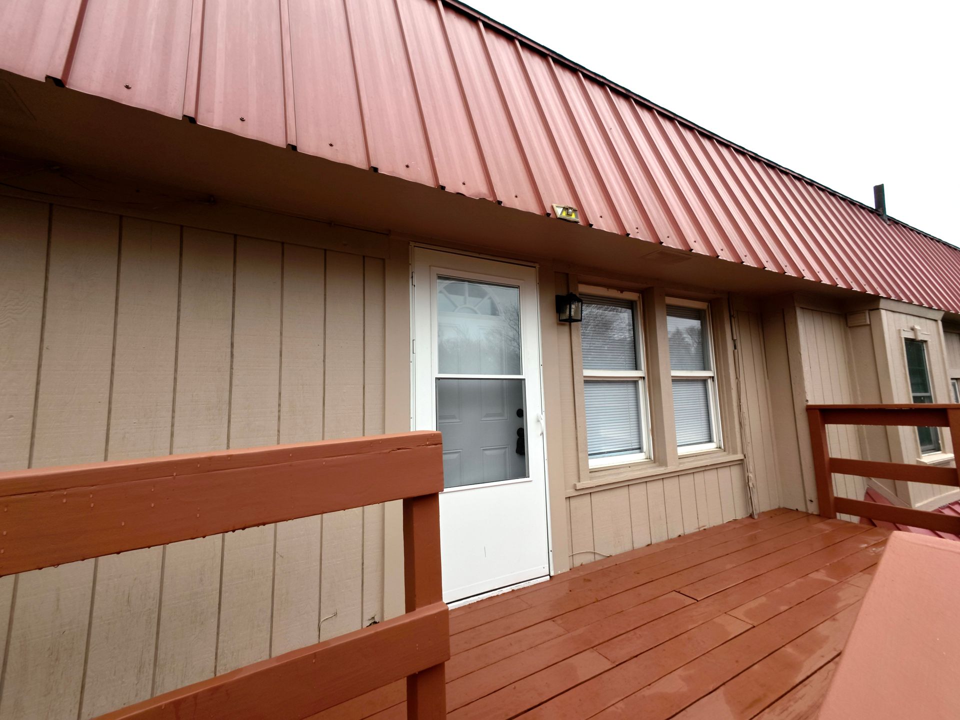 Exterior of a building with a red metal roof, tan siding, white door, and deck with brown railing.