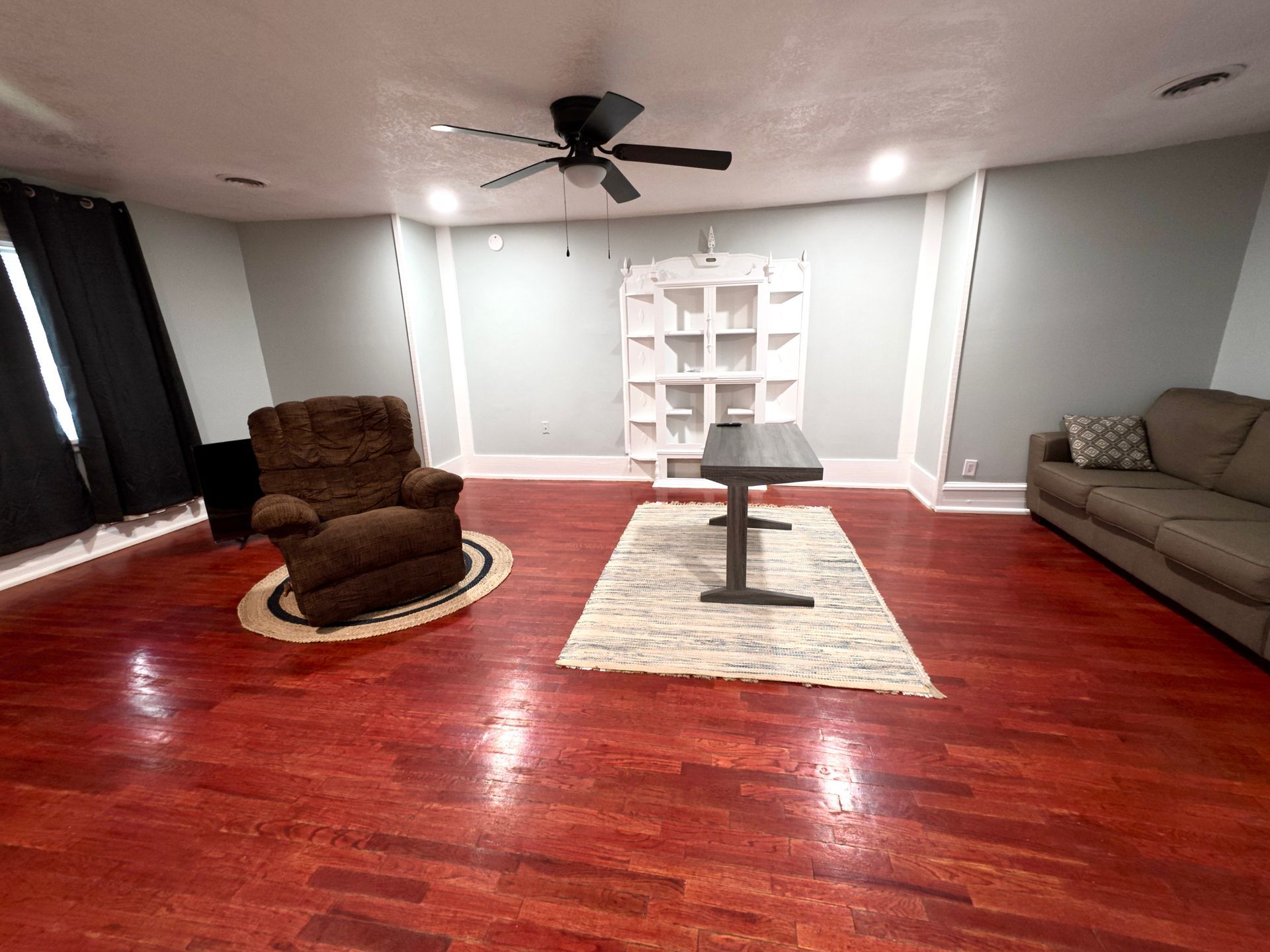 Spacious room with a brown recliner, couch, and wooden floor. A ceiling fan hangs above the furniture.