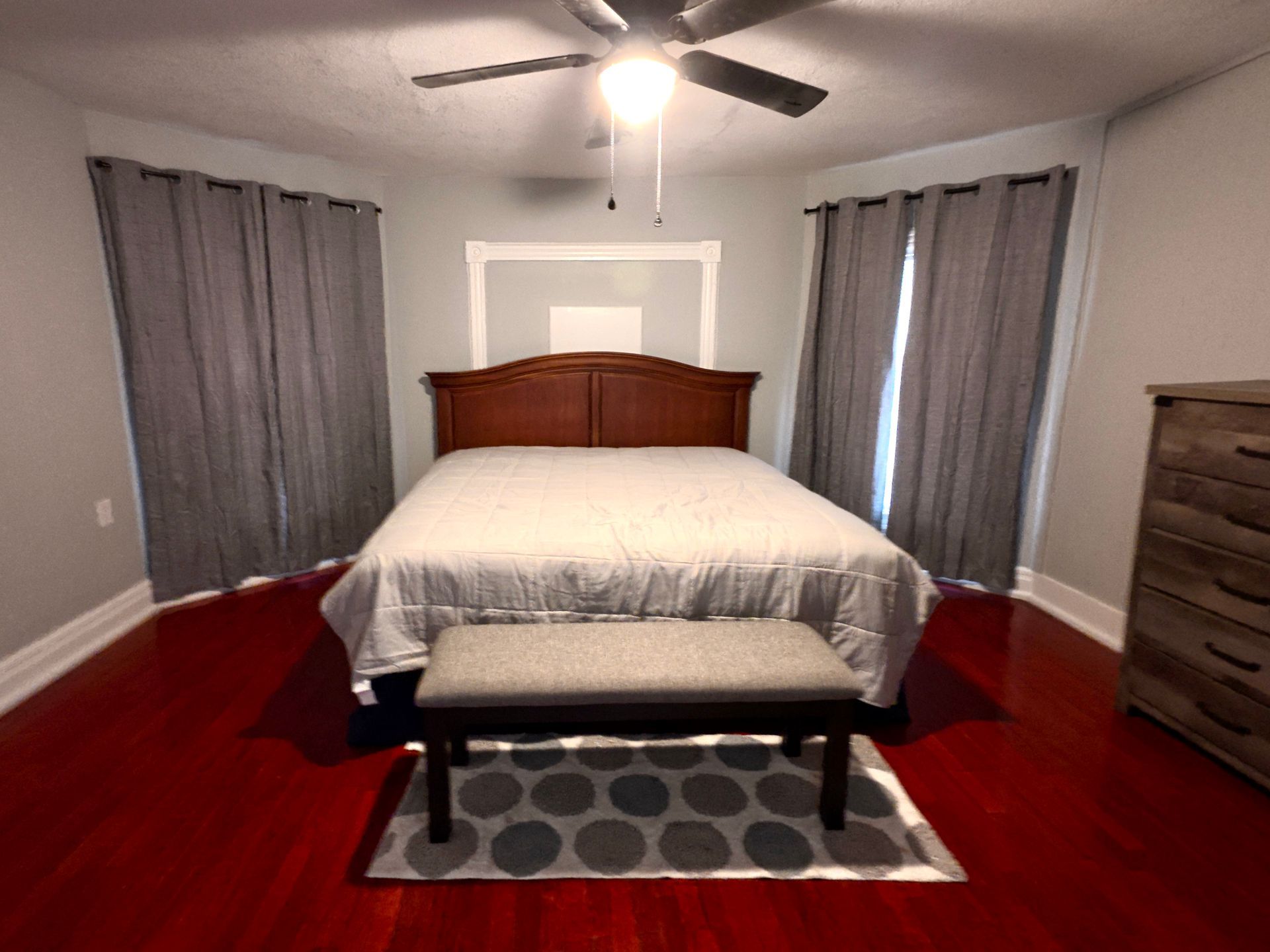 Bedroom with a wooden bed, gray curtains, and dark red floor. A bench sits at the foot of the bed.