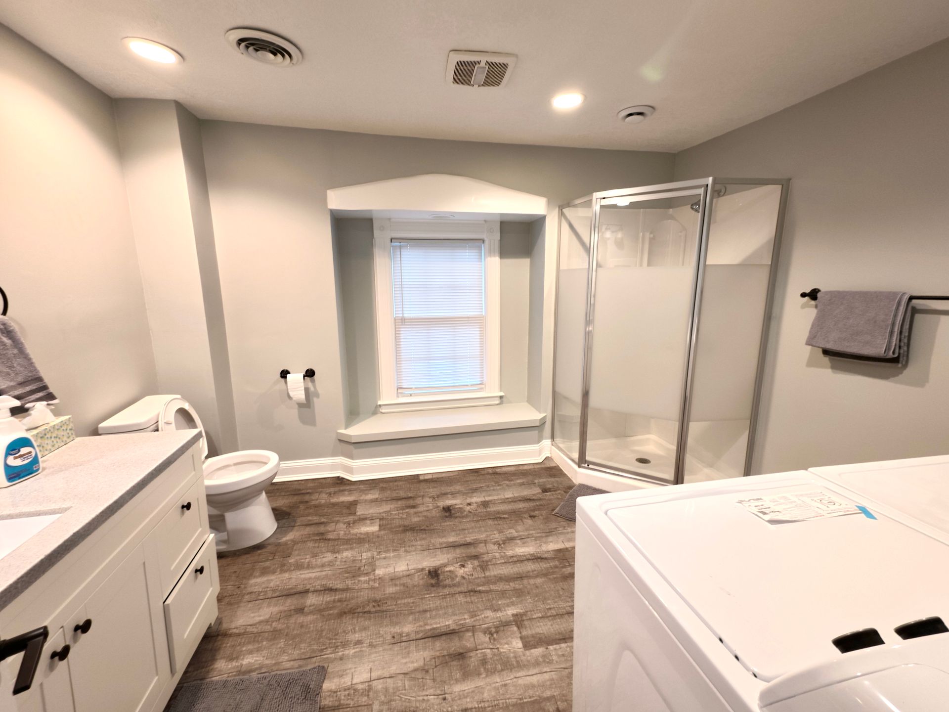 Bathroom with shower, vanity, toilet, and washing machine. Gray walls, wood-look floor, window.