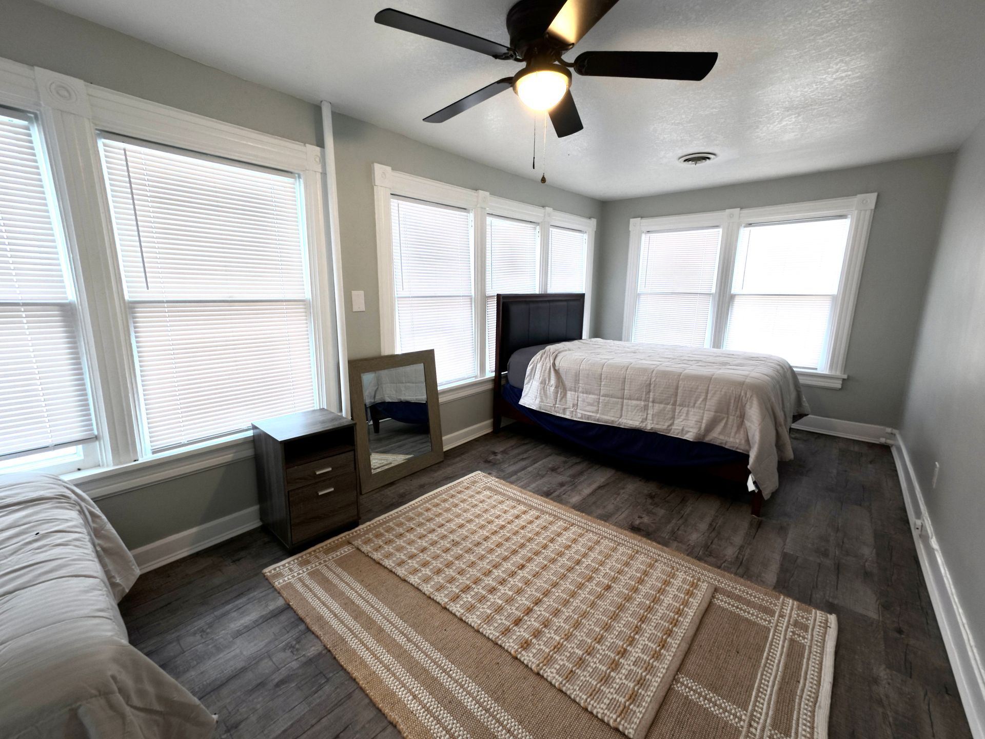 Bedroom with two beds, windows, rug, ceiling fan, and nightstand. Gray walls, wood flooring, and neutral colors.