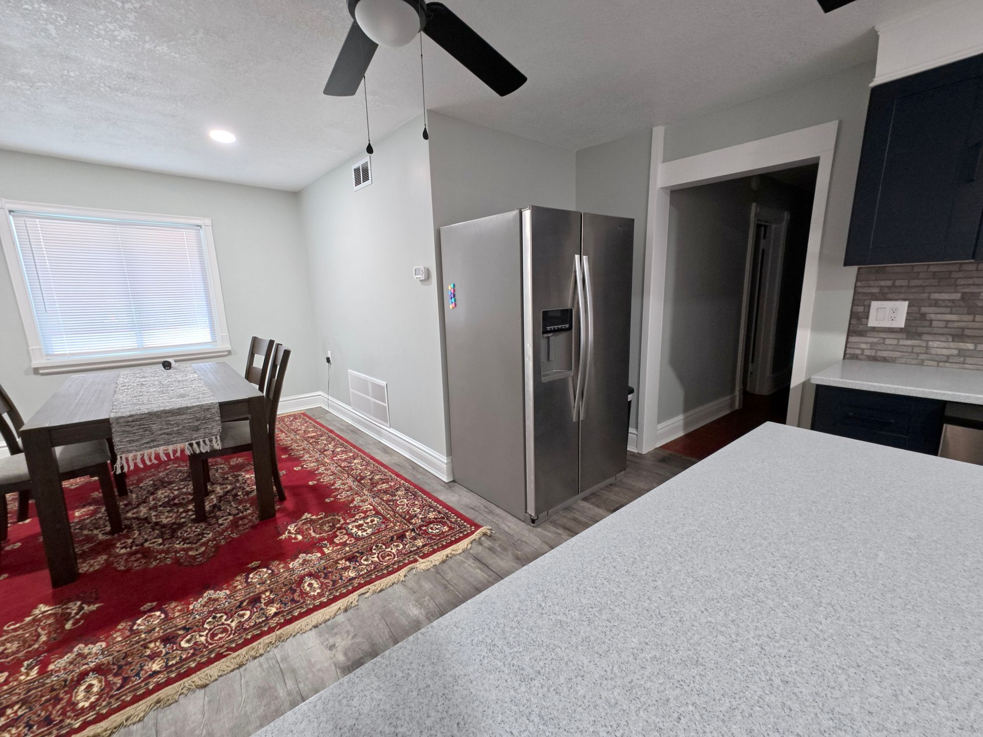 Kitchen with stainless steel refrigerator, dining table with chairs, red rug, and gray walls.