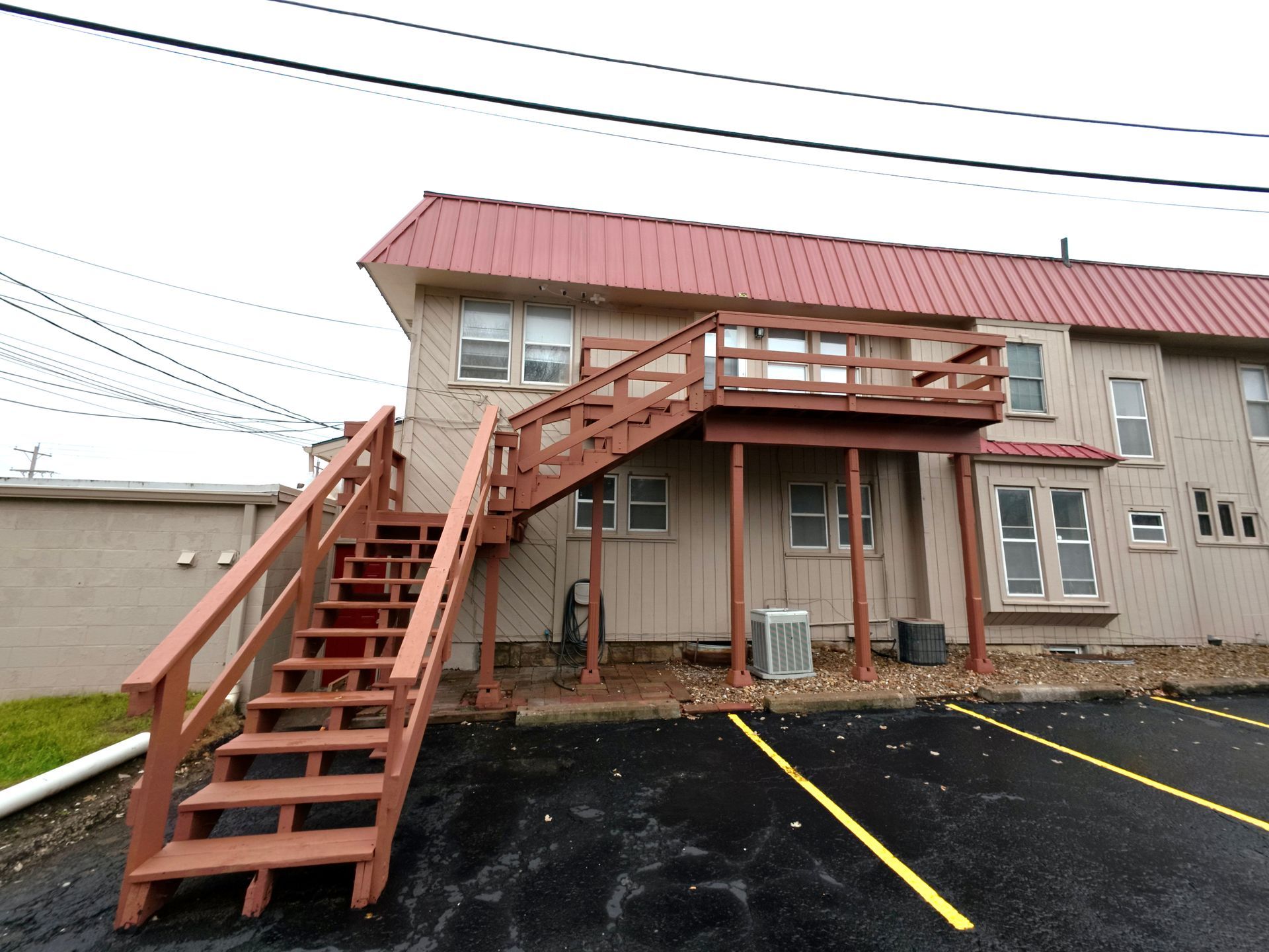 Exterior of a two-story building with red stairs leading to a deck; a red roof and parked cars in front.