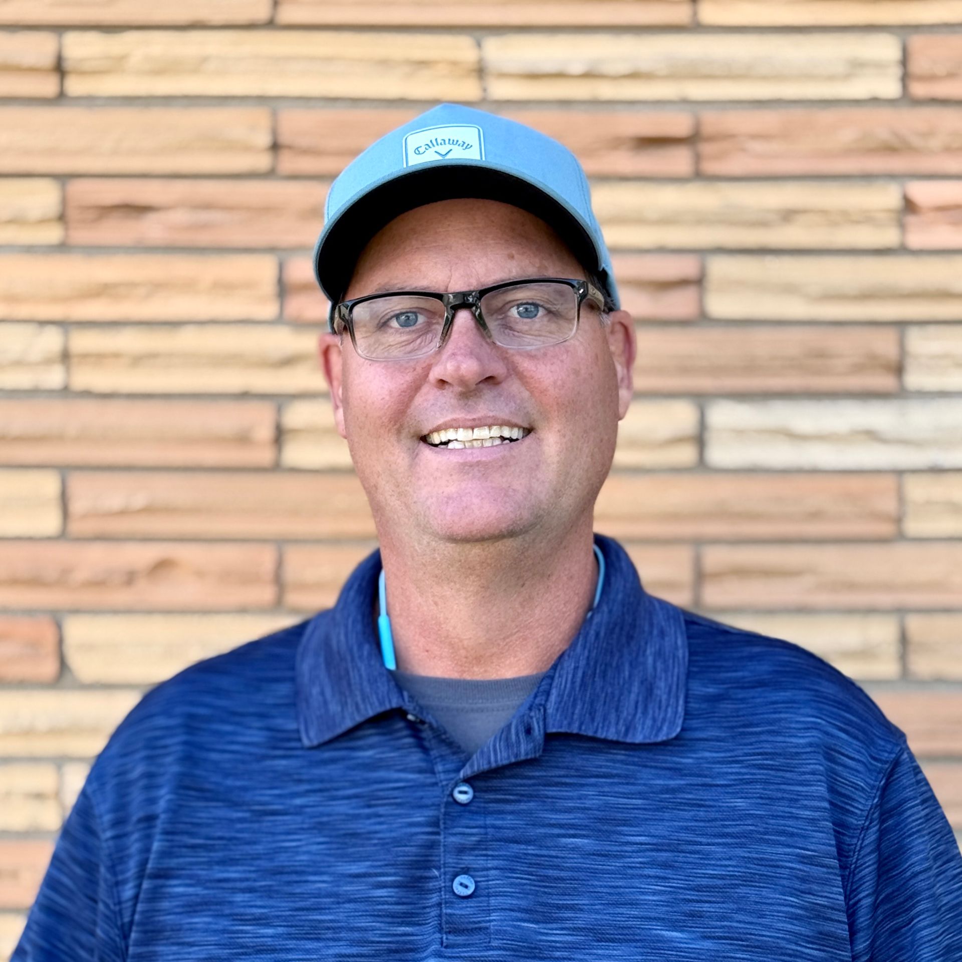 Man wearing blue hat and polo smiling in front of a brick wall.