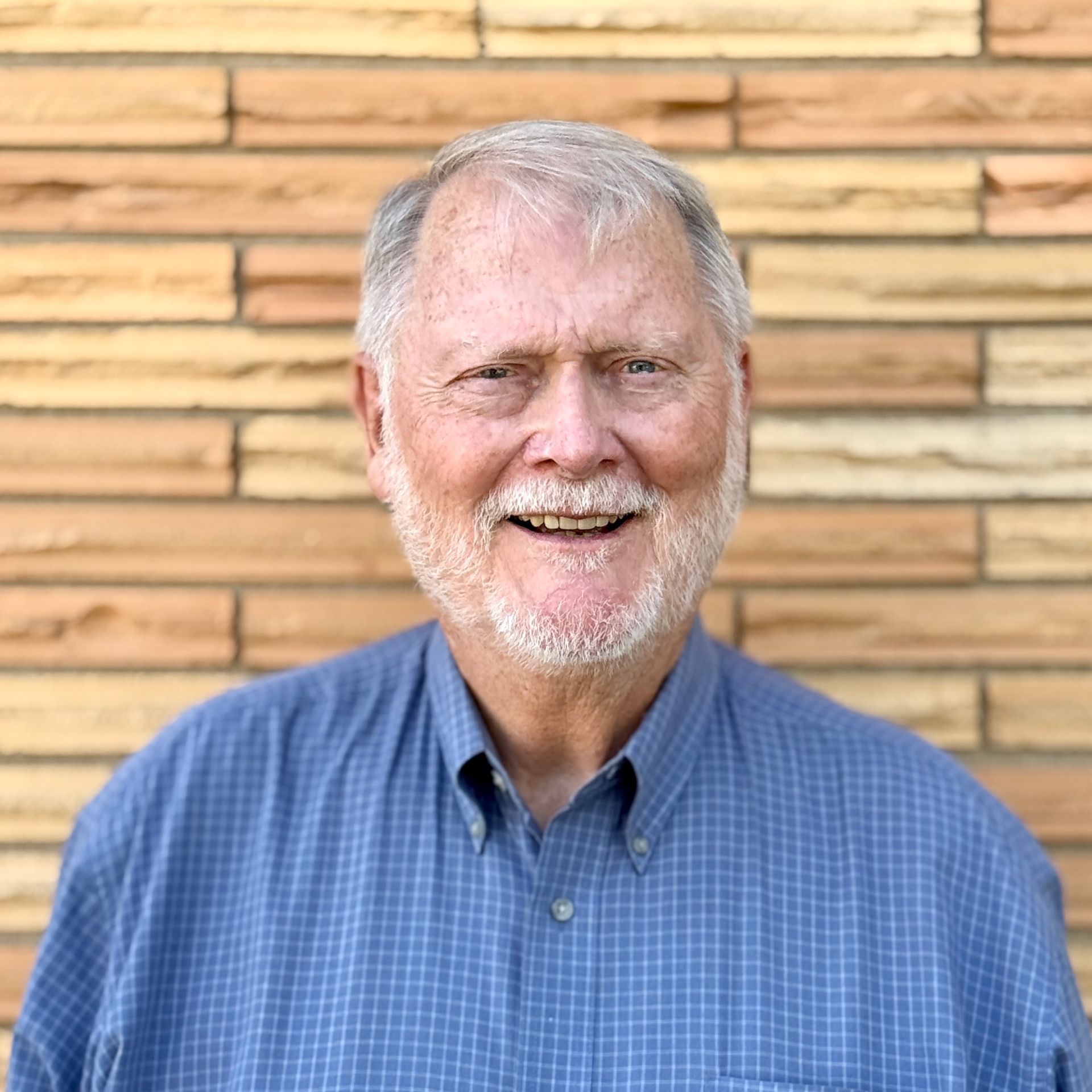 Smiling man with gray hair and beard, wearing a blue button-down shirt, in front of a brick wall.