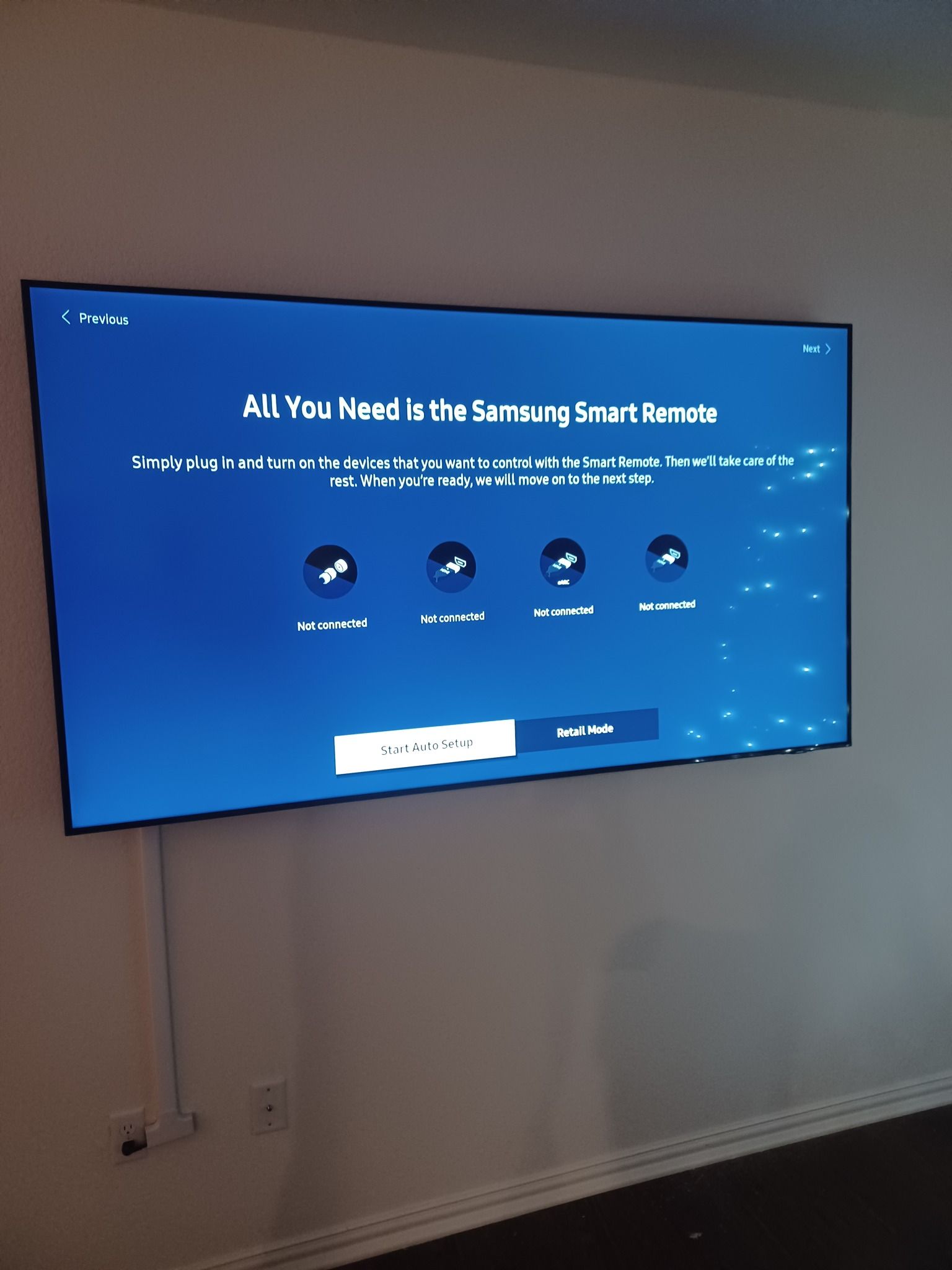 A wall-mounted Samsung TV displaying a blue setup screen with remote control prompts. White wall, black baseboard.