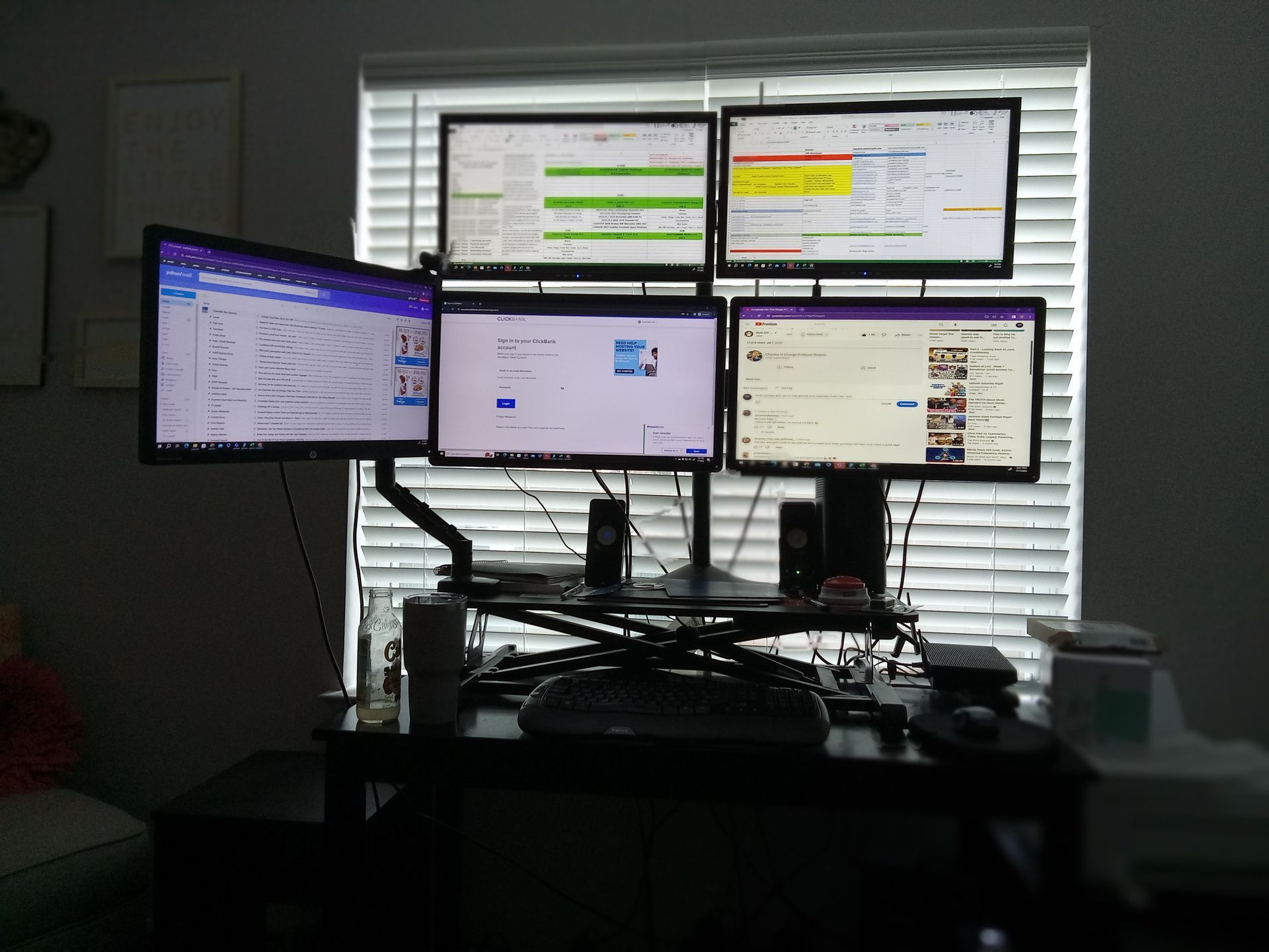 A desk with five computer monitors displaying various applications; black desk and monitor arms.