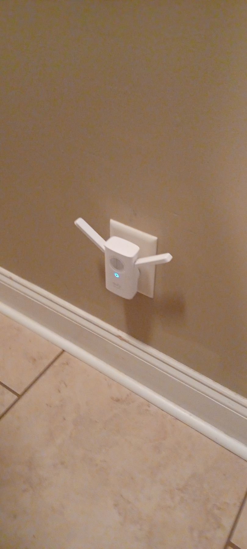 White WiFi extender plugged into a wall outlet on a tan wall; tiles and a baseboard are in view.