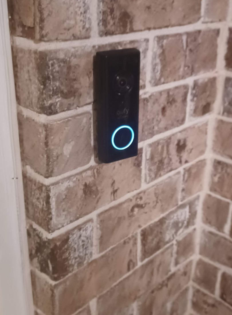 Black doorbell with a blue illuminated ring mounted on a brick wall.