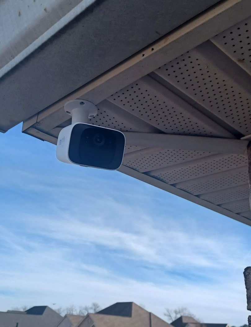 White security camera mounted on the underside of a roof overhang, against a blue sky with clouds.