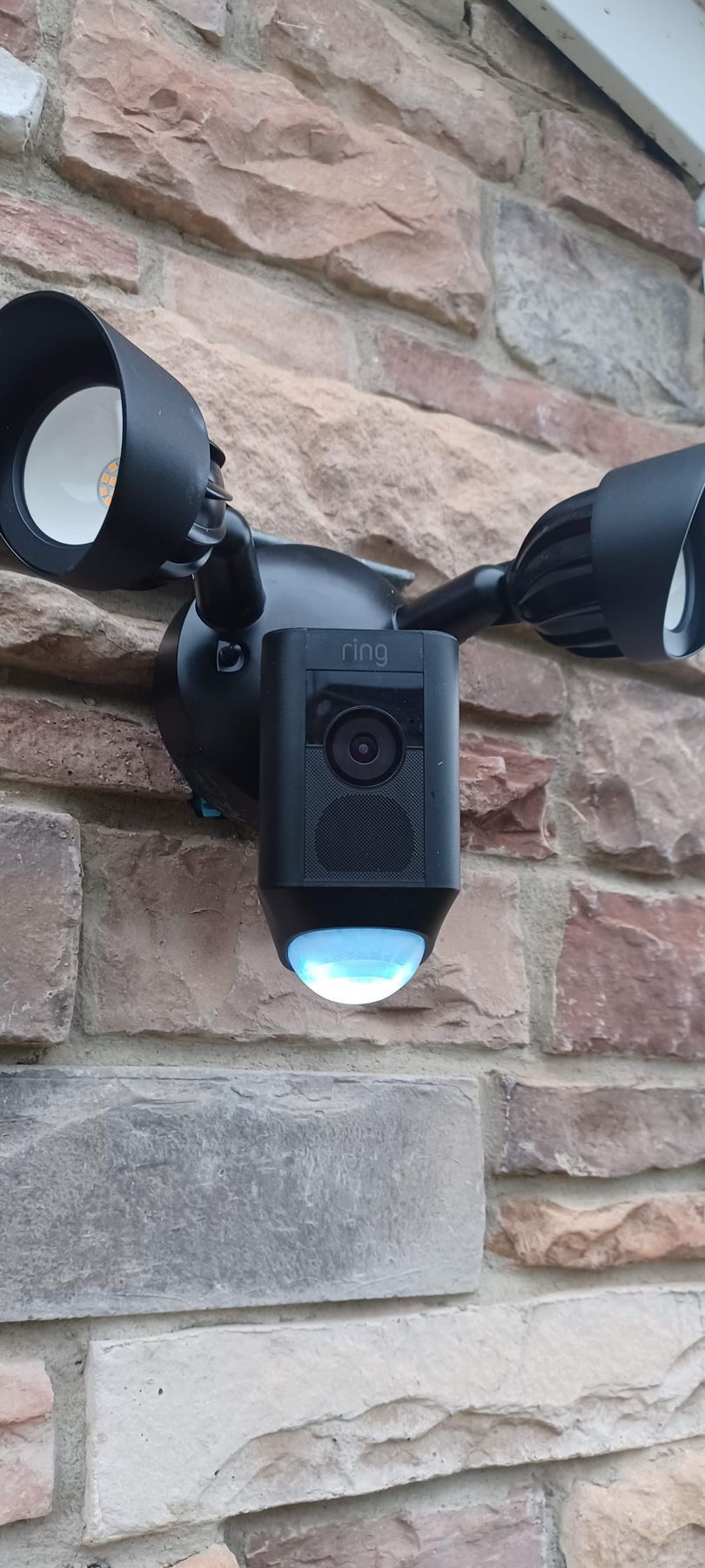 Black security floodlight with camera mounted on a brick wall.