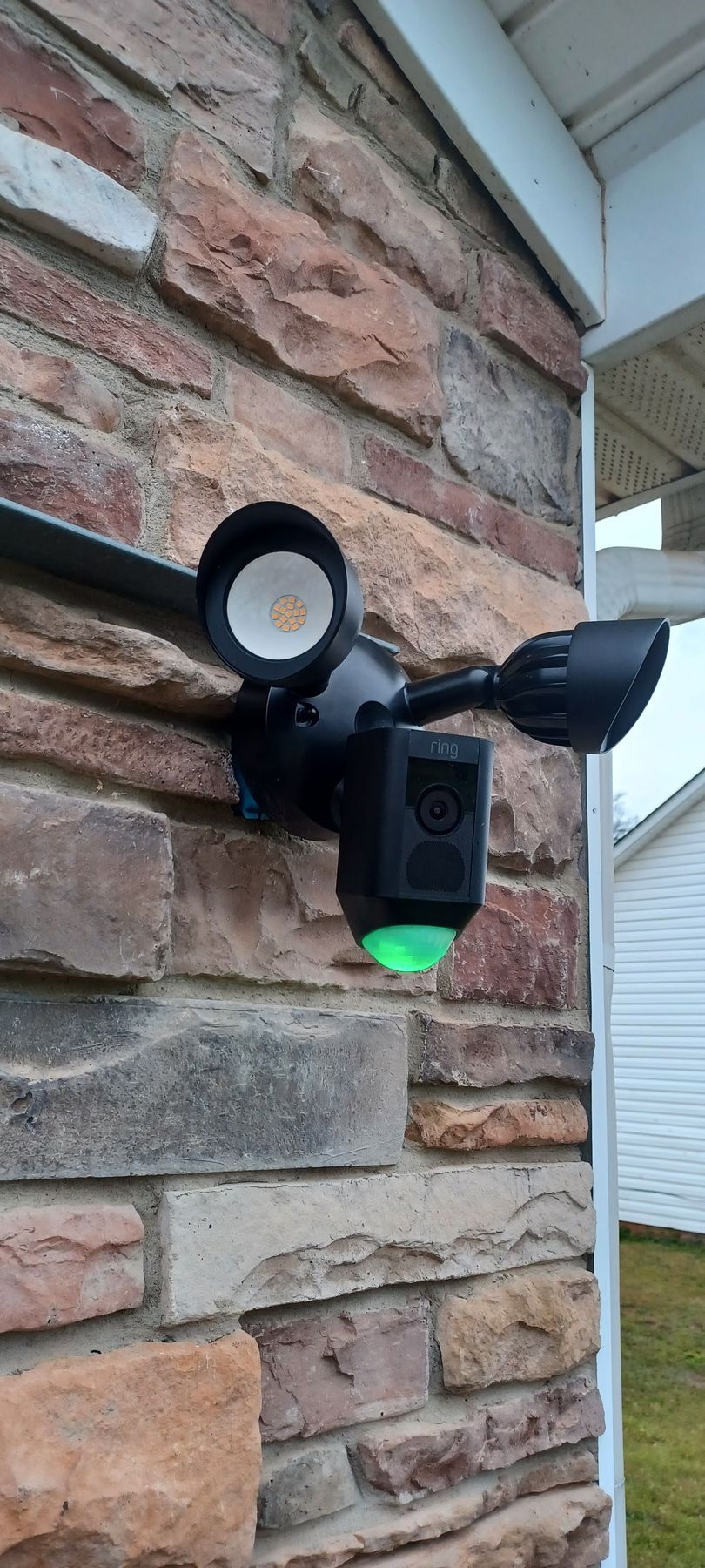 Black security camera with lights attached to a brick wall; a green light is illuminated.