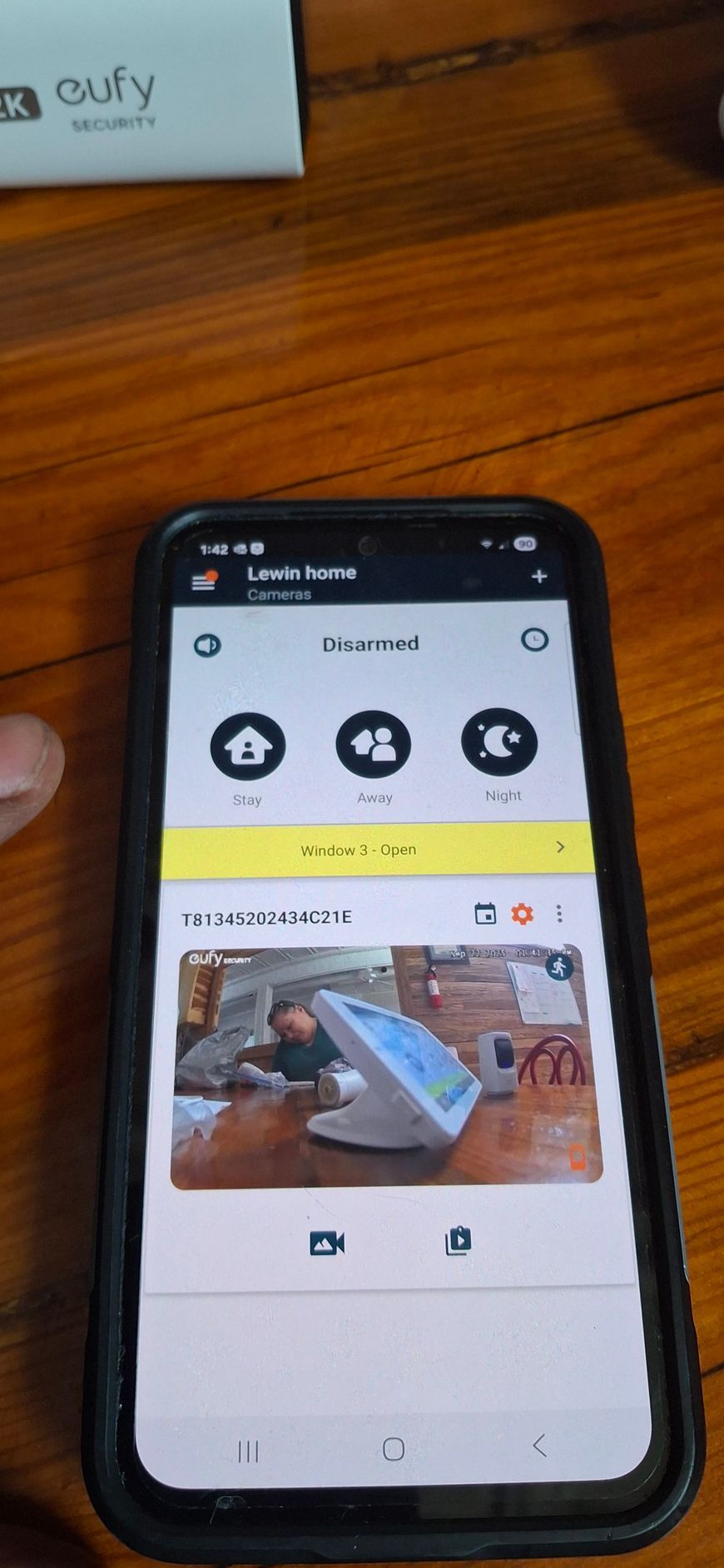 A phone displaying a live video feed of a person, likely from a security camera, sitting on a wooden table.