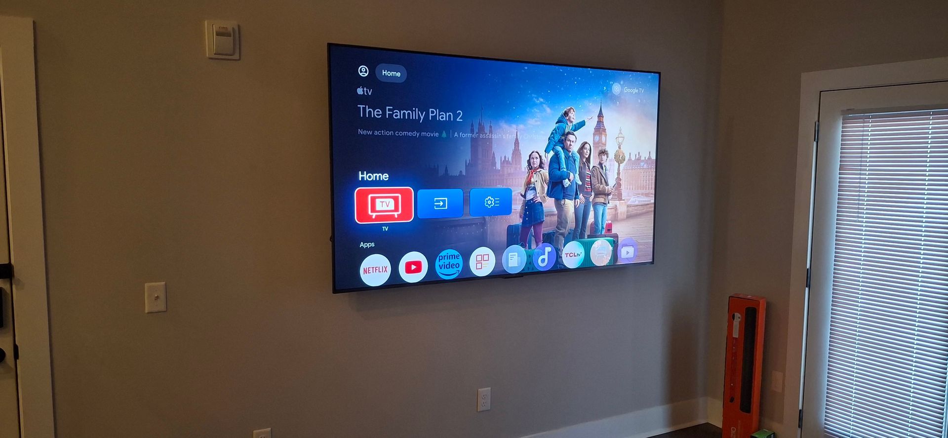 A large TV mounted on a wall. The screen displays a colorful menu. A door and window are in the background.