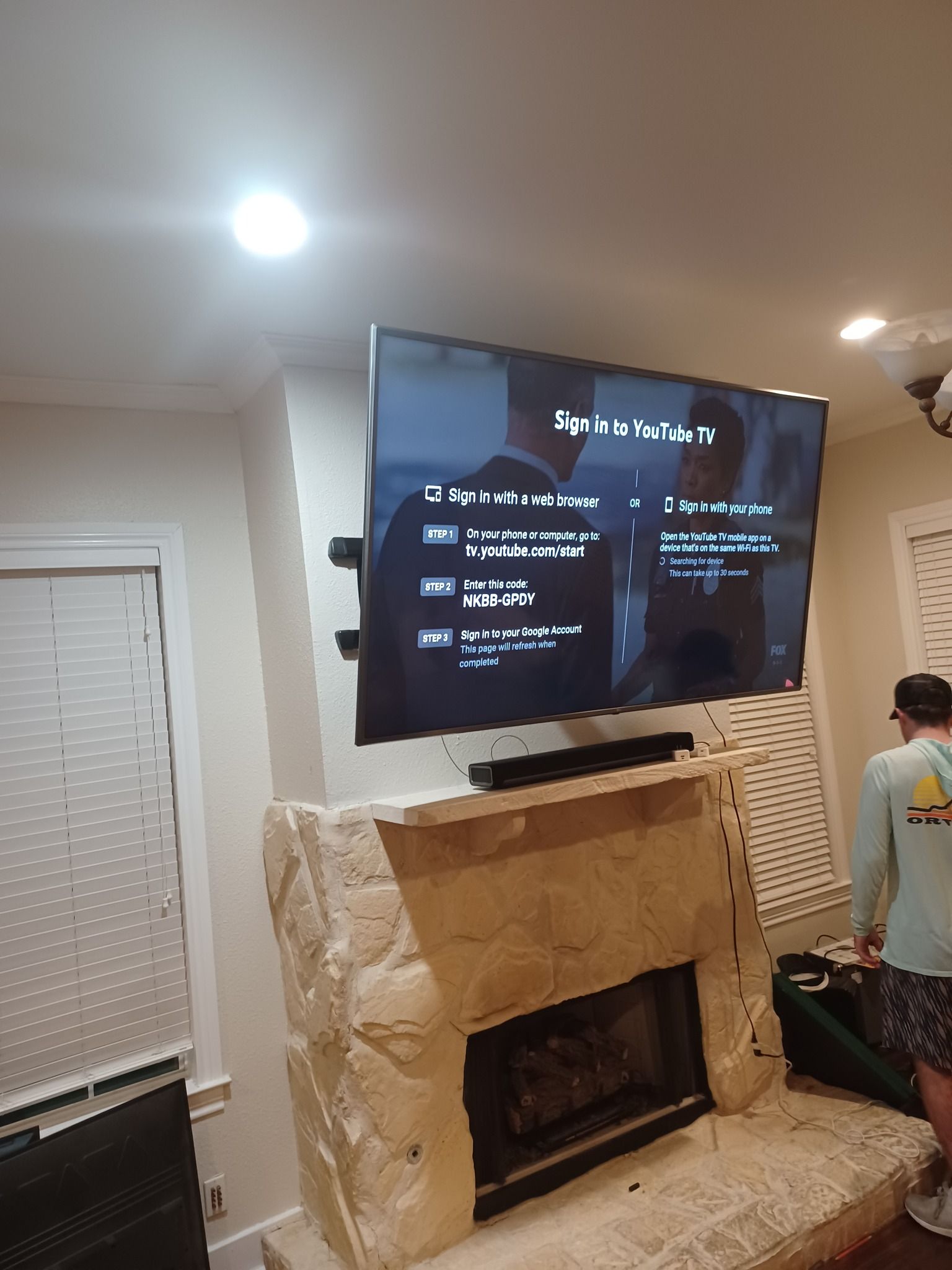 TV mounted above a stone fireplace. A person carries a case in the room. The TV displays a streaming service interface.