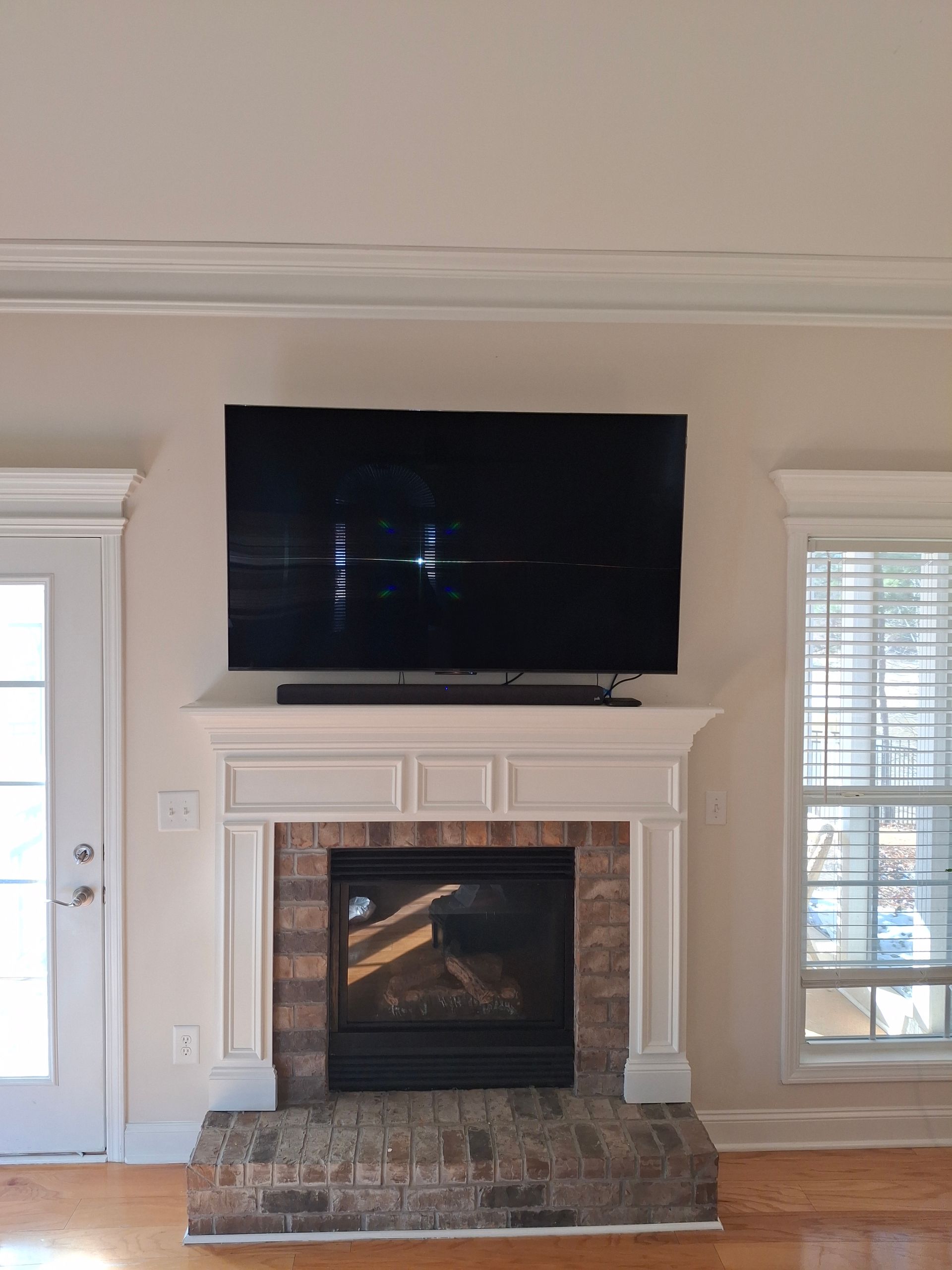 Flat-screen TV mounted above fireplace with white mantel and brick hearth, in a room with a door and window.