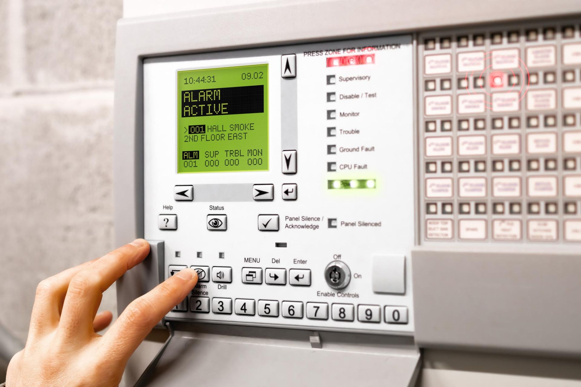 Hand pressing a button on a gray fire alarm control panel with a green display, numerous buttons, and red lights.