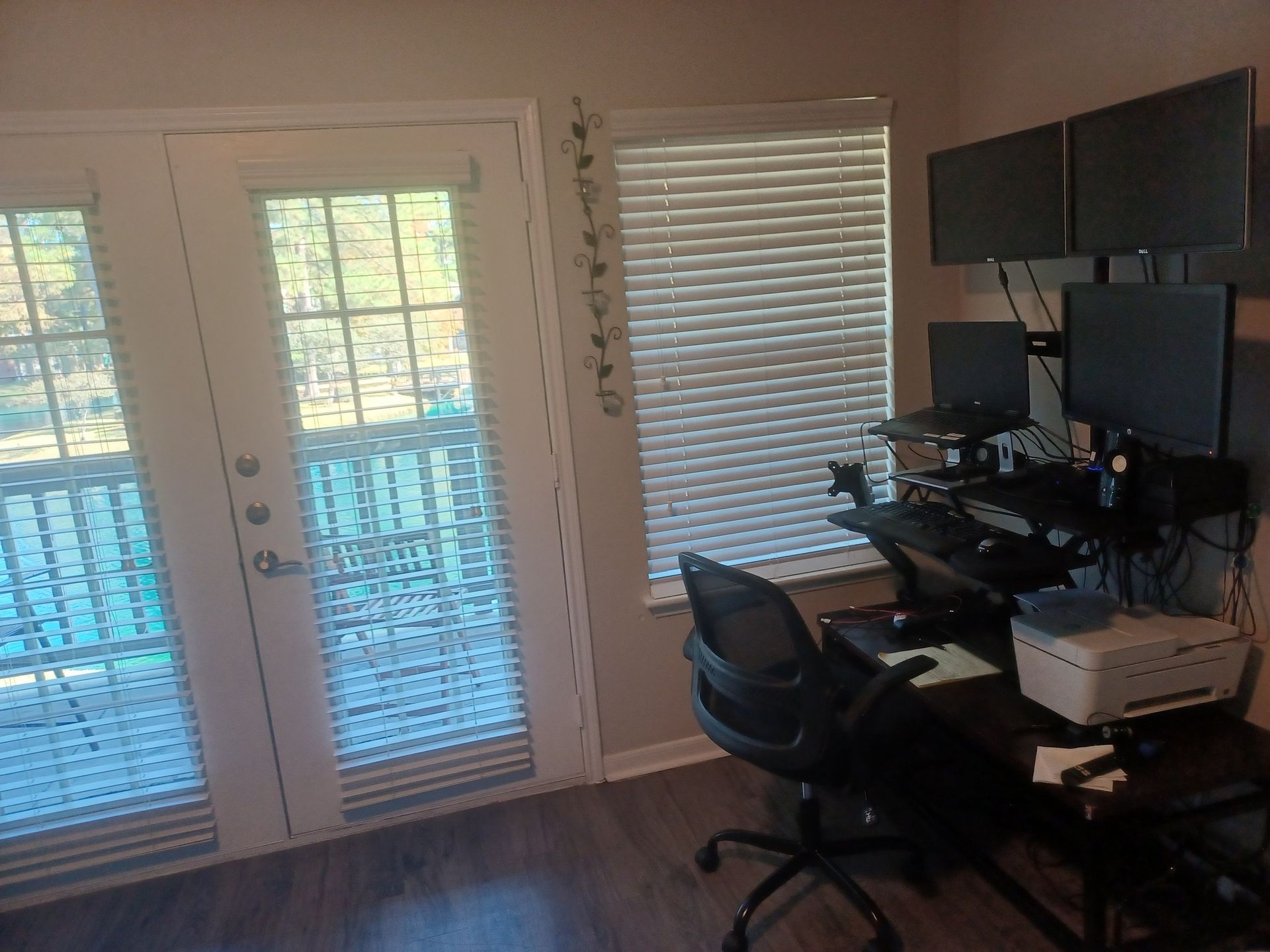 Office setup with desk, multiple monitors, chair, and a door to a balcony with blinds.