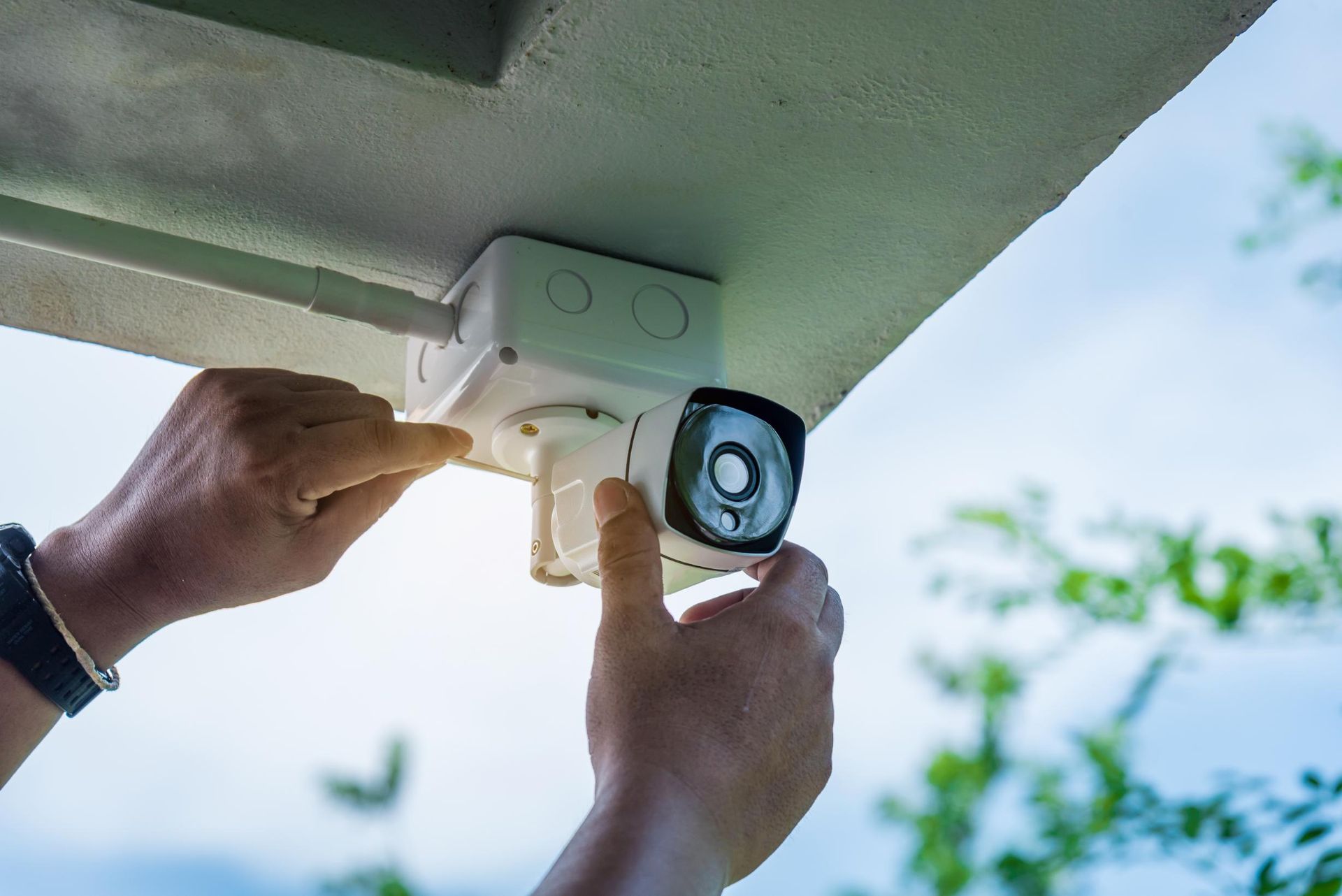 Person installing a security camera under a white overhang with green foliage in the background.