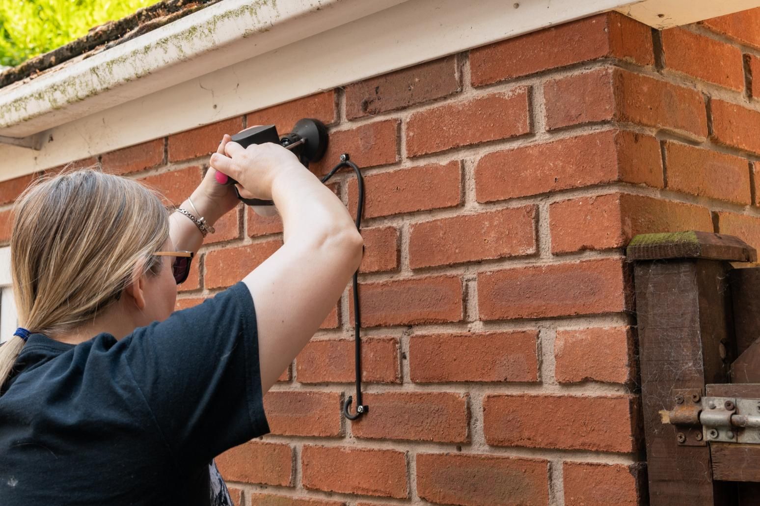 Person attaching a black object to a red brick wall outdoors.