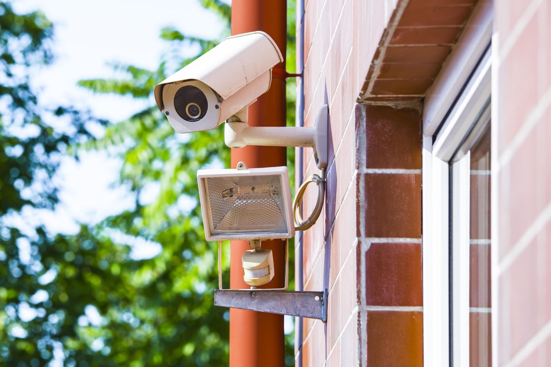 Security camera and floodlight mounted on a brick wall next to a window; leafy green trees in background.