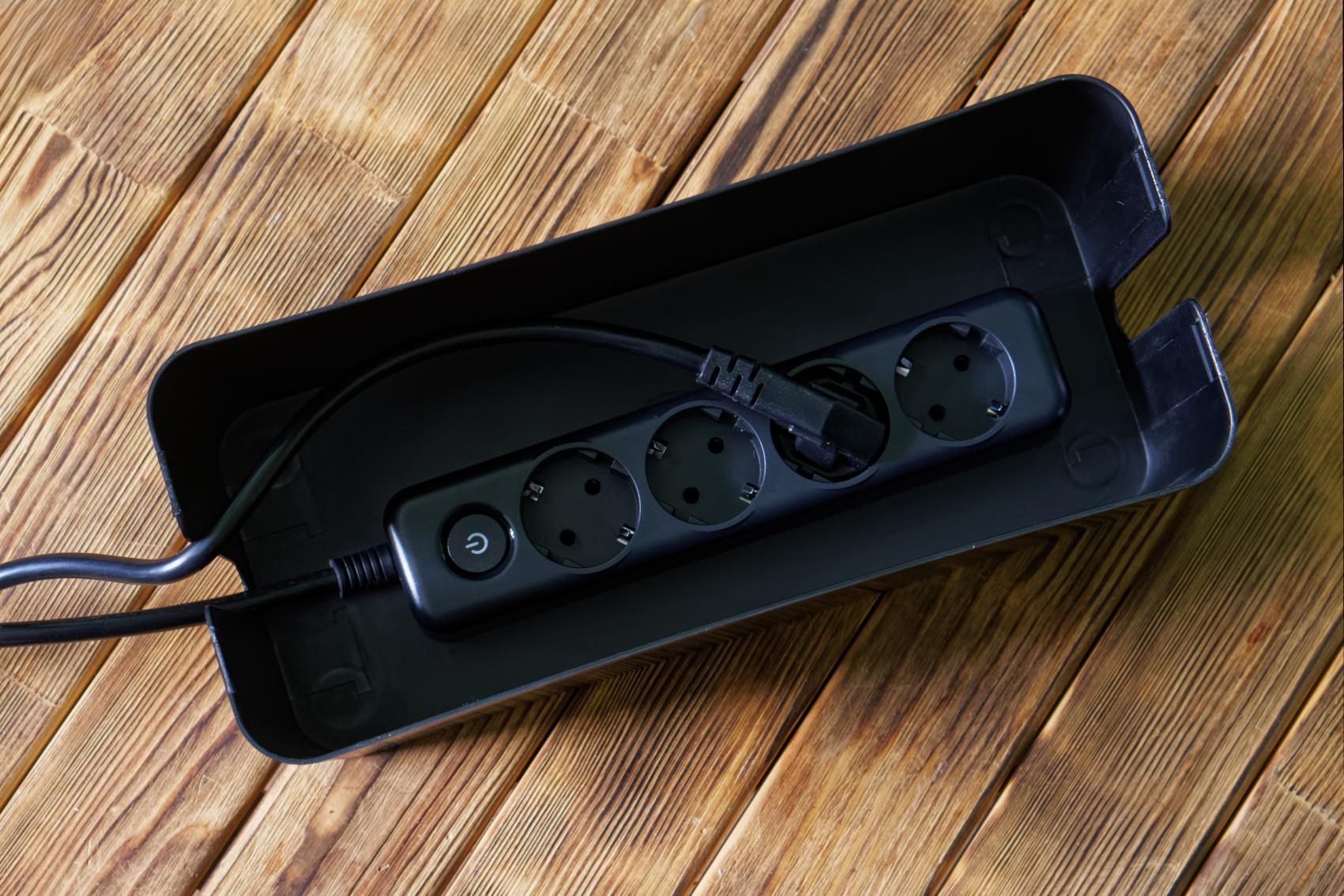 Black power strip inside a black cable organizer on a wooden surface.