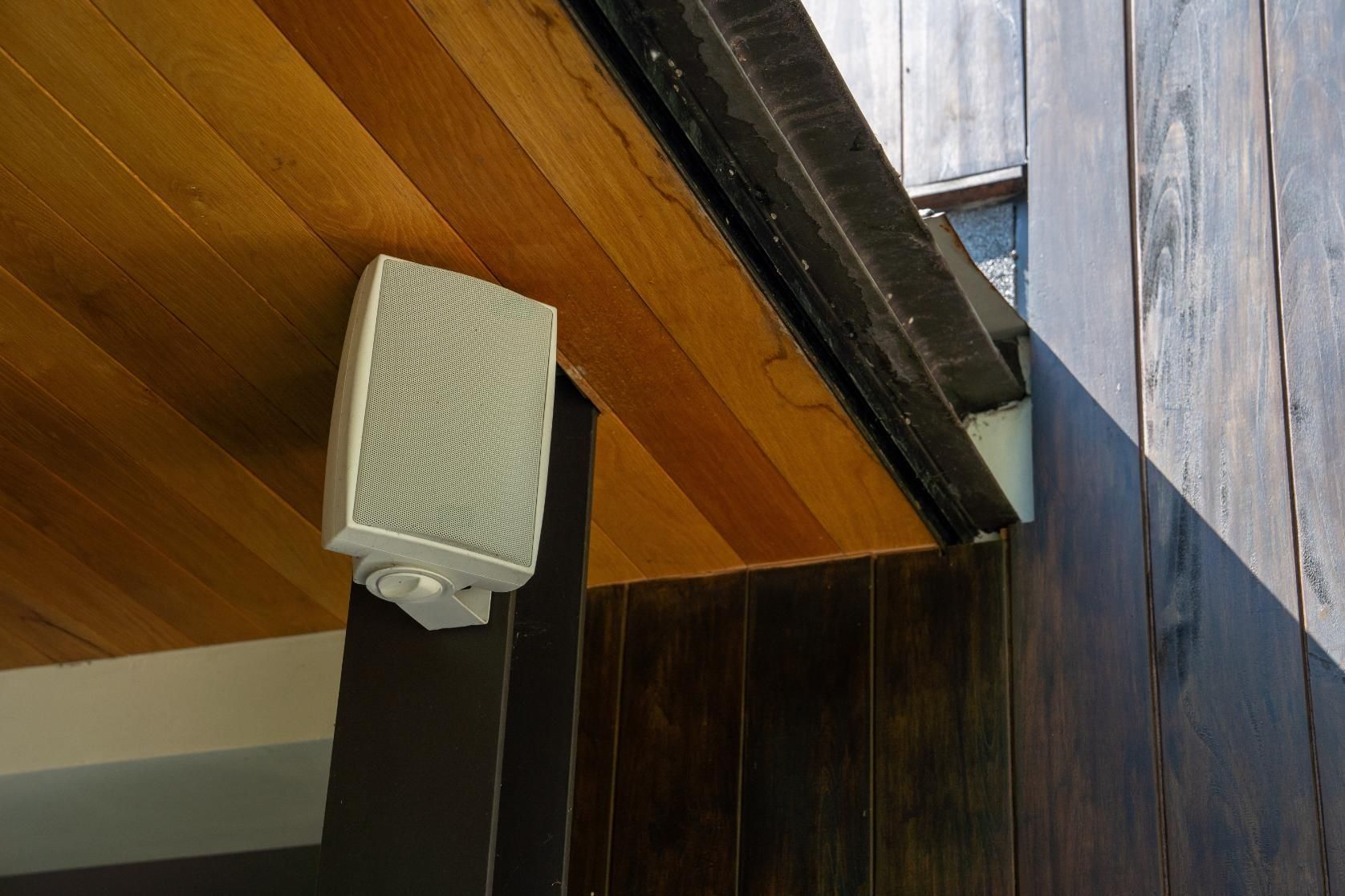 White outdoor speaker mounted on a wood ceiling, next to a brown wooden exterior wall.