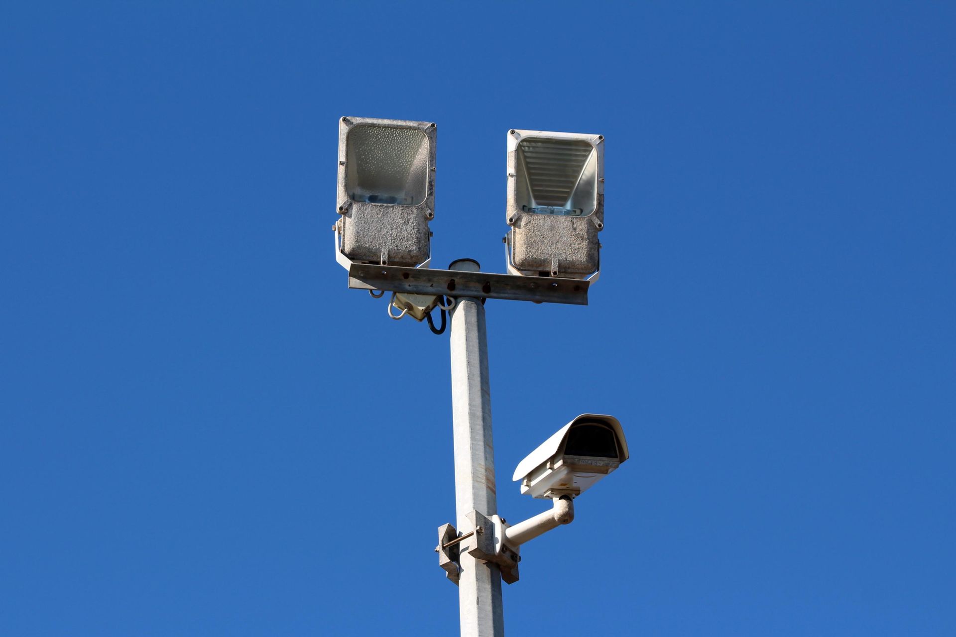 Light pole with two floodlights and a security camera against a blue sky.