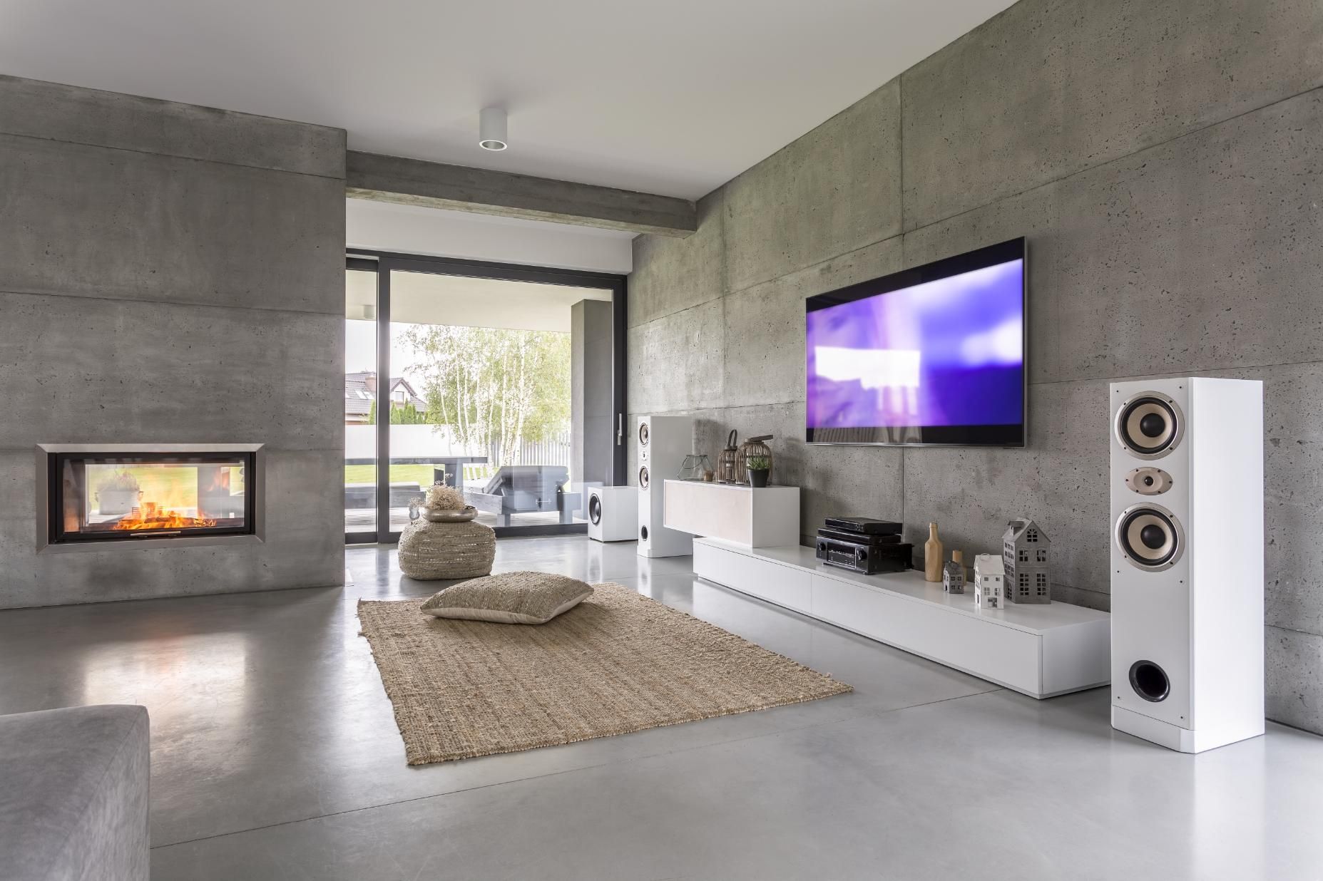 Modern living room with concrete walls, fireplace, large TV, white speakers, and a rug.