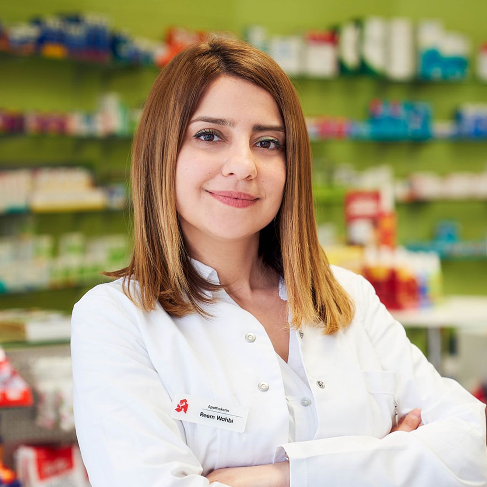 Eine Frau in einem weißen Laborkittel steht mit verschränkten Armen in einer Apotheke.
