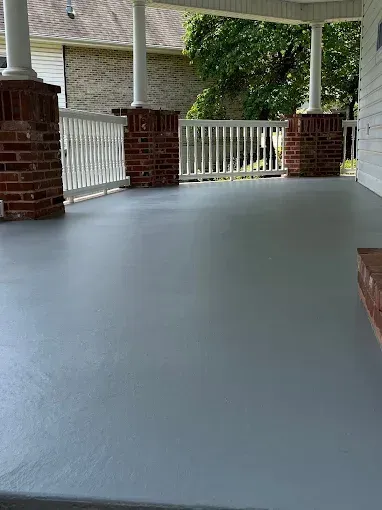 Gray-painted porch with brick and white pillars, white railing, and a glimpse of greenery.