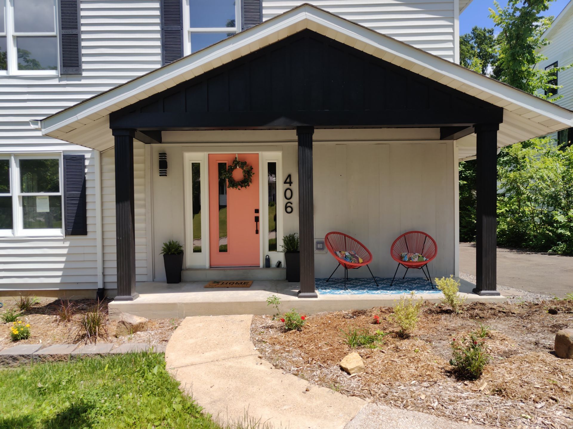 Front of a house with peach door and black accents. Porch has two red chairs and a blue rug.