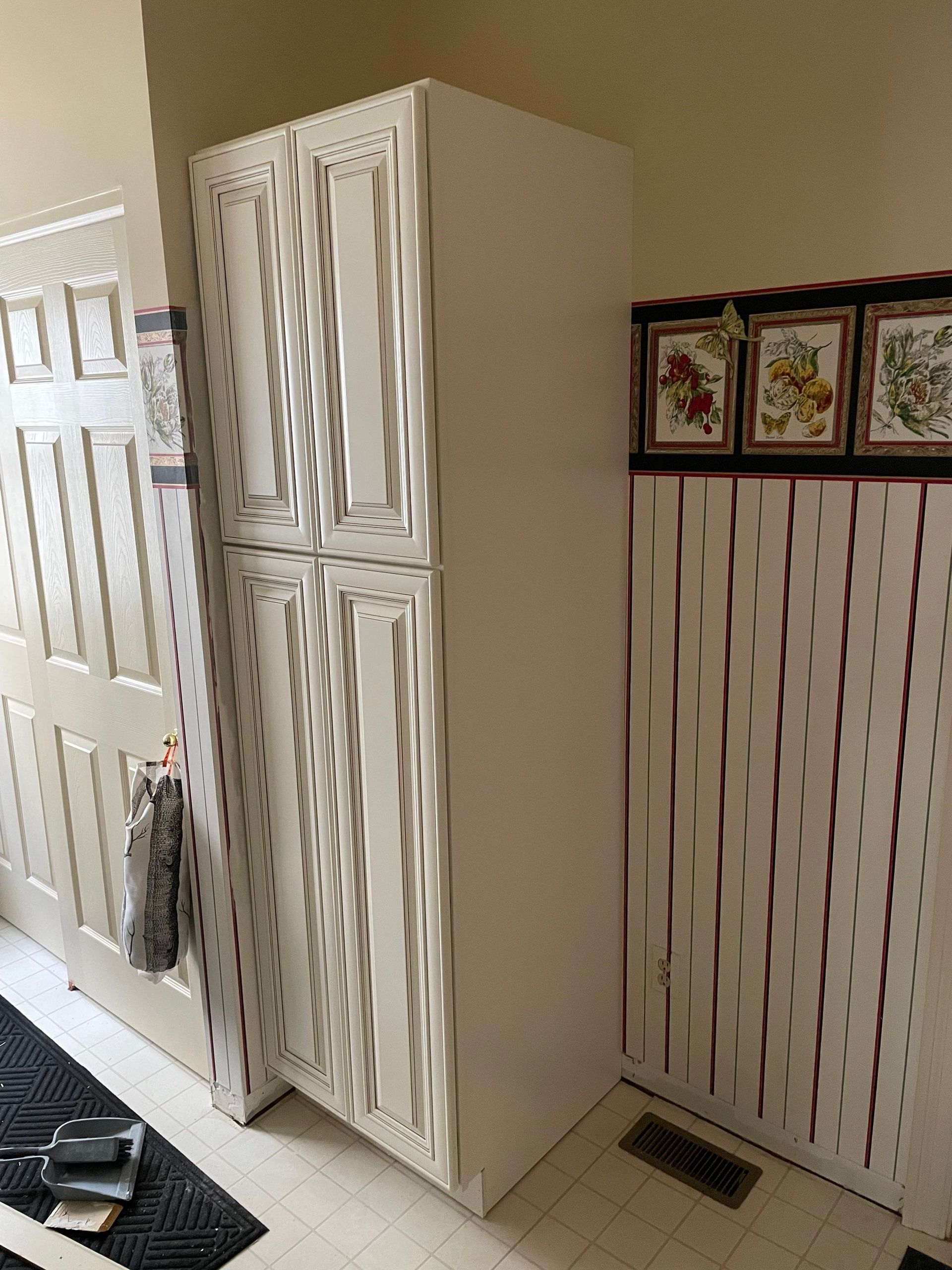 Tall white pantry cabinet next to a patterned wall and a doorway with a rug and door.