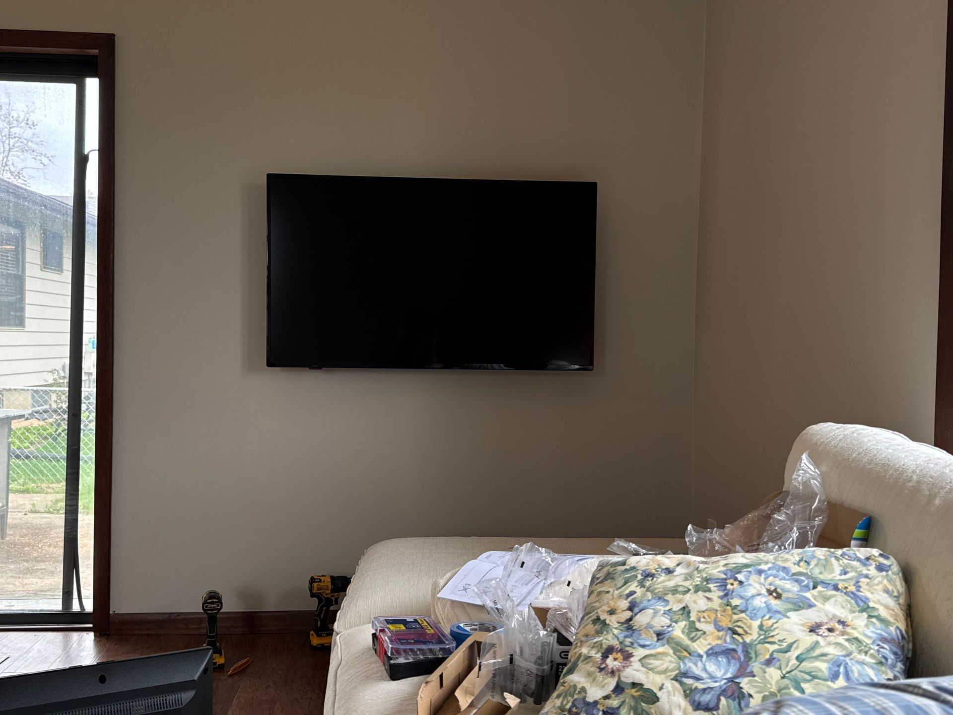 TV mounted on a beige wall above a couch with floral cushions near a window.