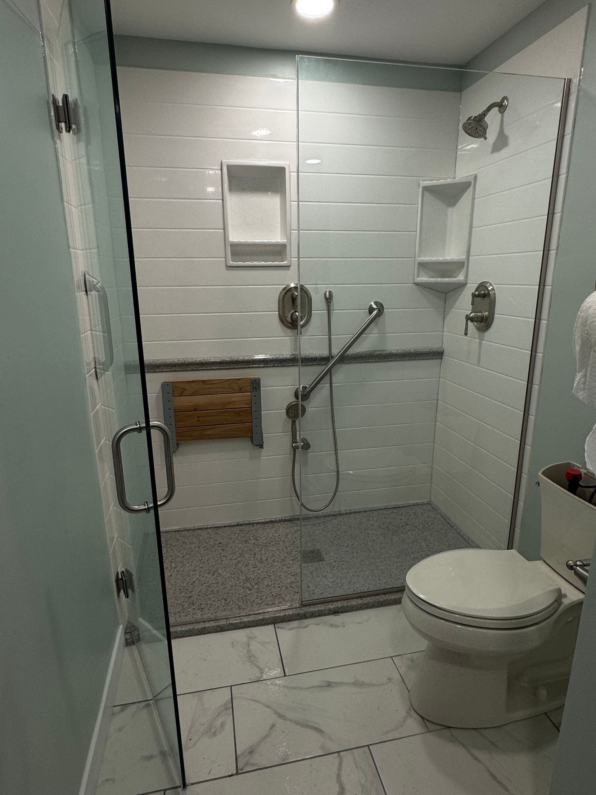 Modern bathroom with glass shower, white tiles, wood bench, and toilet.