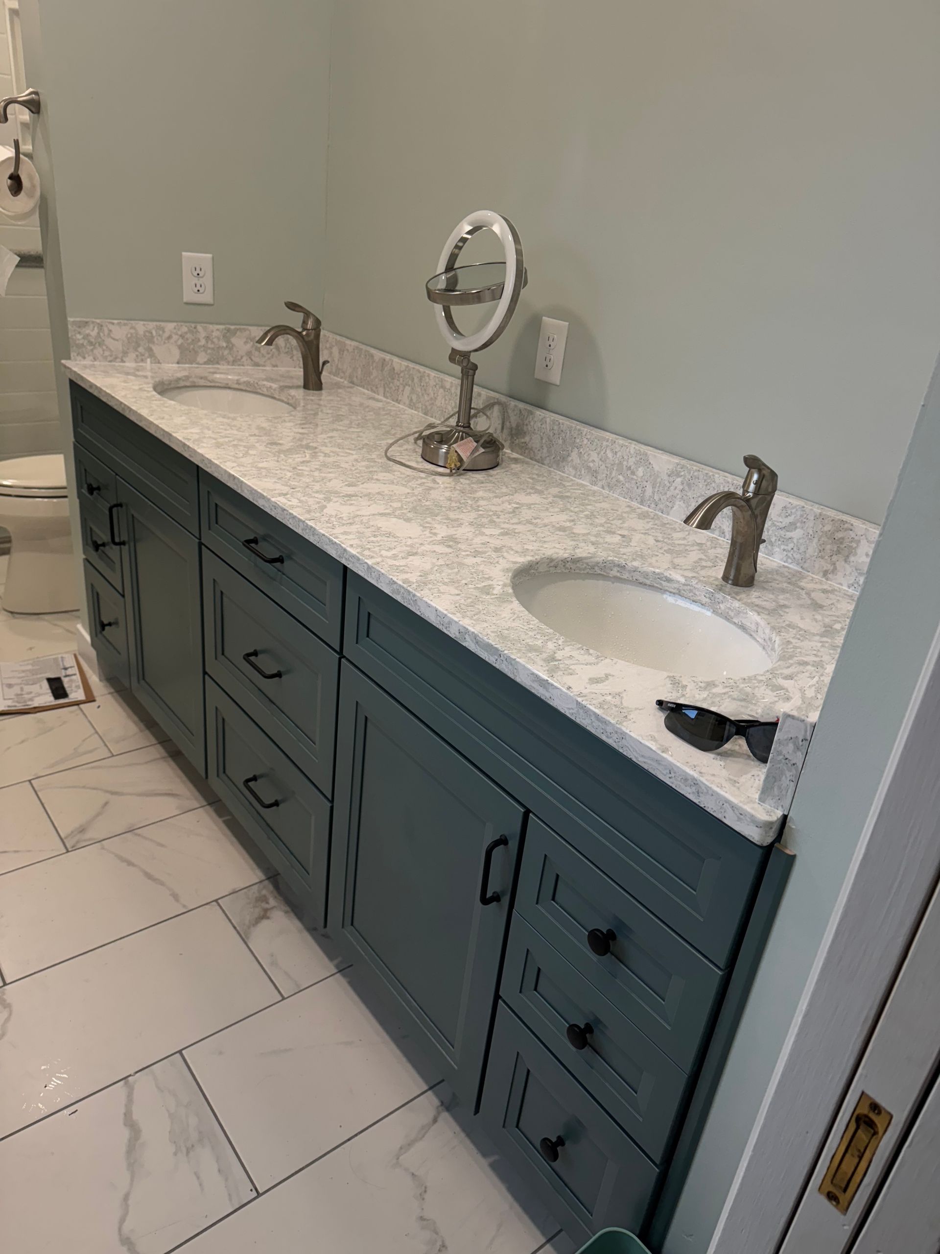 Blue bathroom vanity with white countertop, two sinks, and a round mirror.