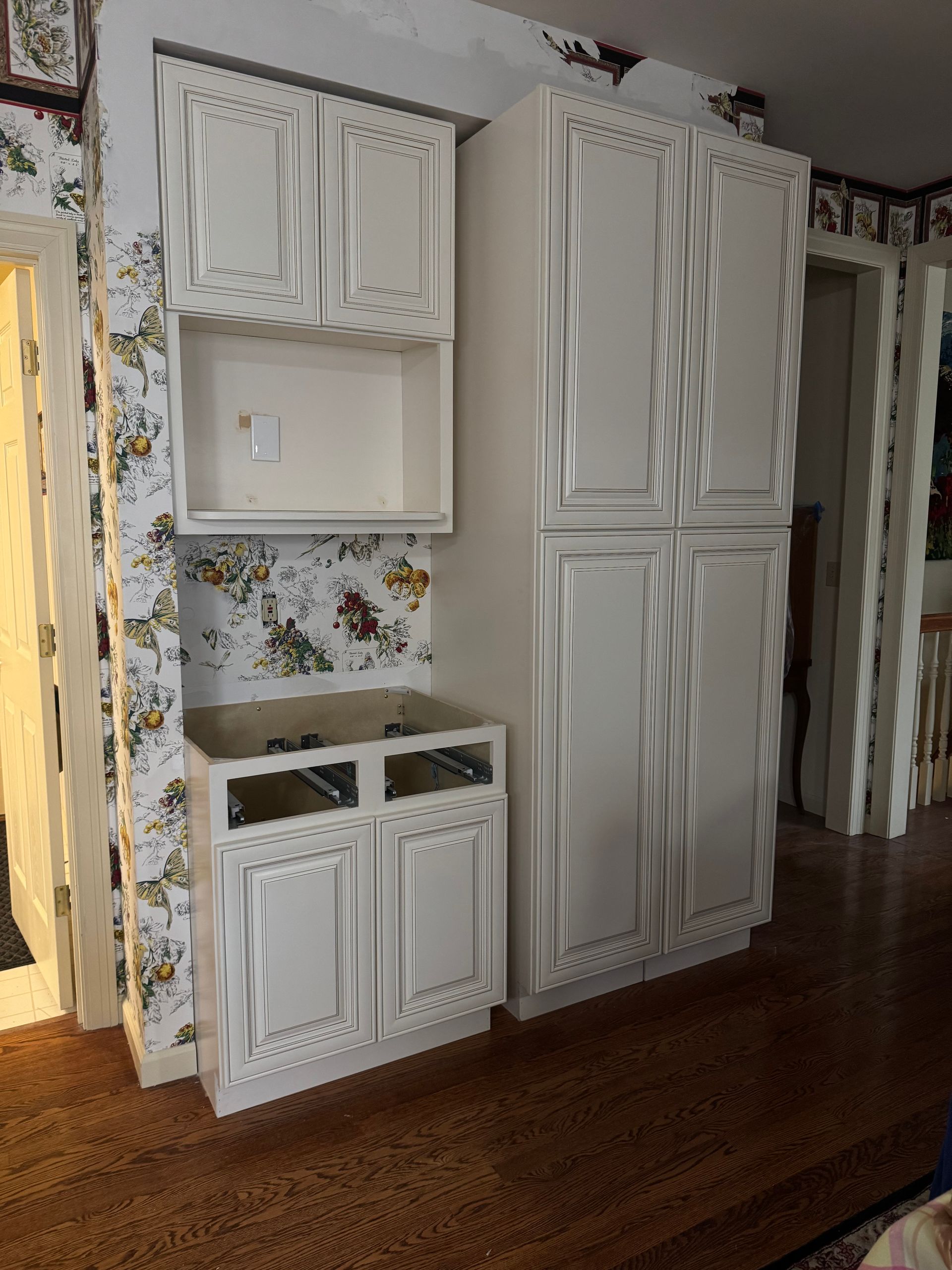 White kitchen cabinets, some with wavy designs, installed against floral wallpaper, on wood floor.