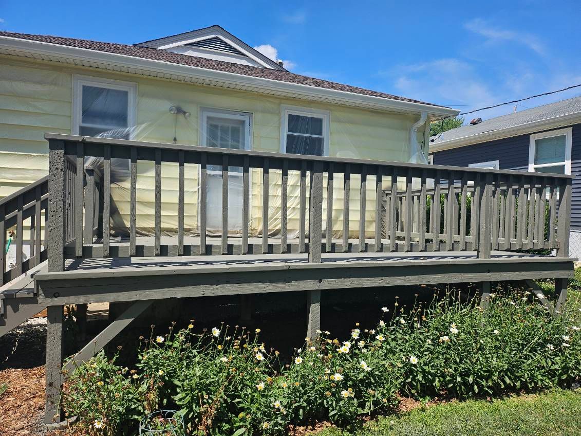 Wooden deck attached to a yellow house, with railing, surrounded by green plants and a blue sky.