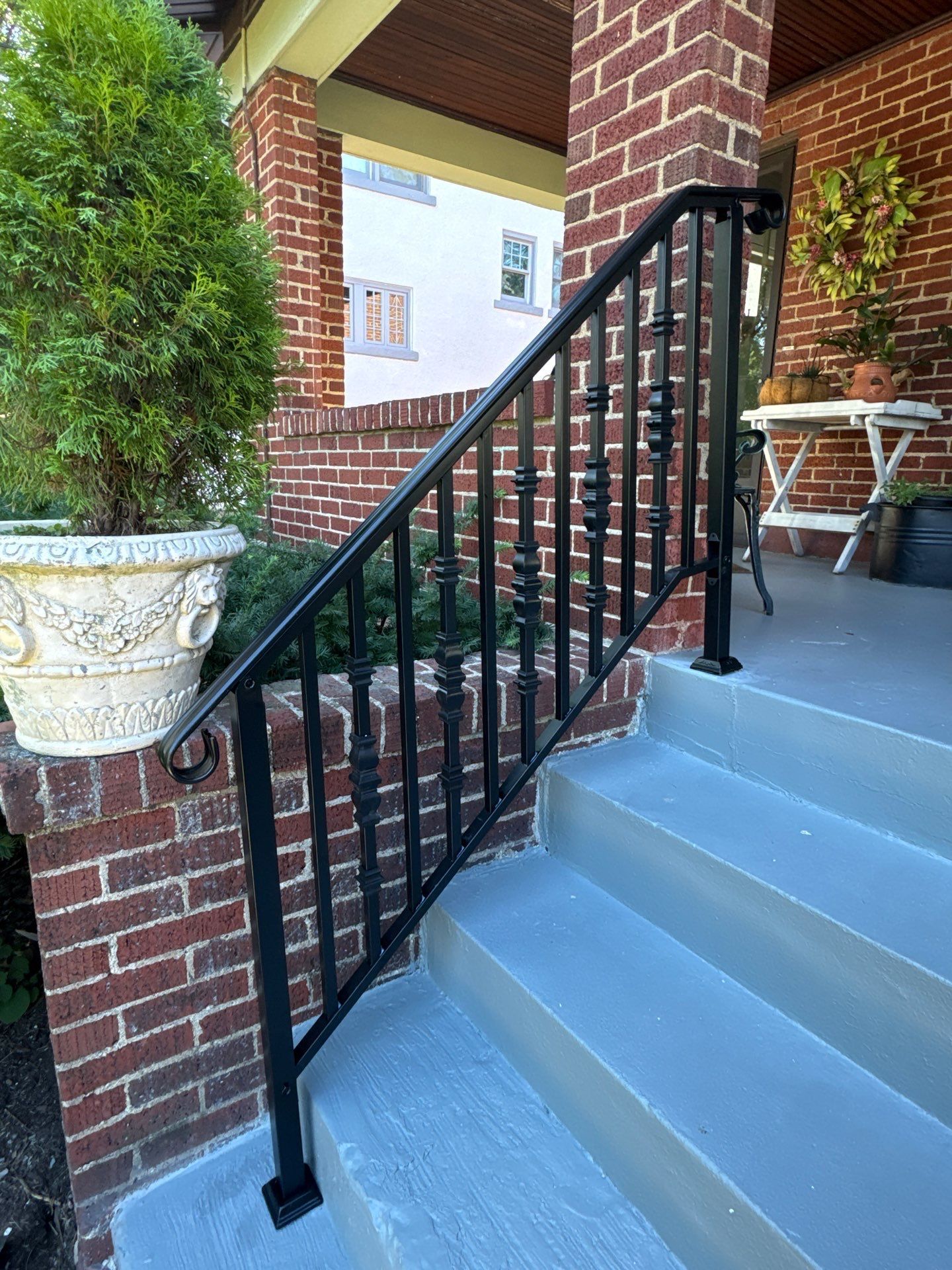 Black metal railing on brick steps. Gray painted concrete. Green plants in pots.