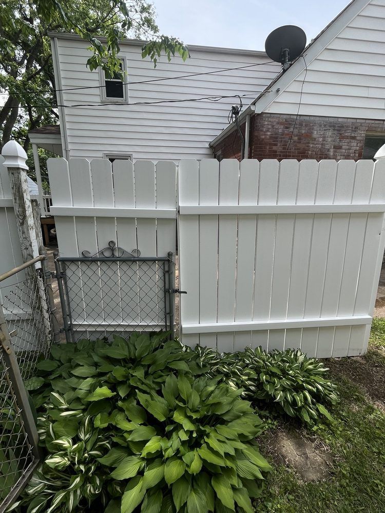 White picket fence with a gate, next to a house, and hosta plants in the foreground.