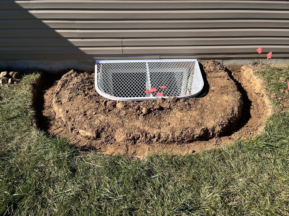 Dirt piled around a window well with a metal grate, next to a building with brown siding.