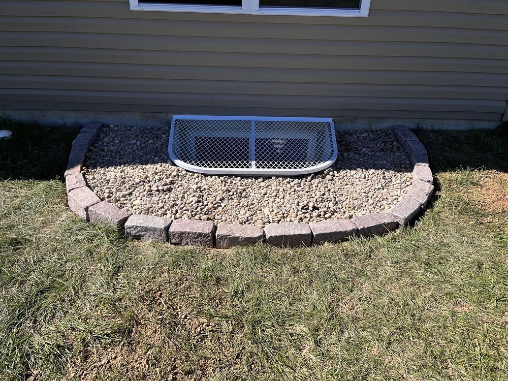 Window well with gravel and brick border, covered by a metal grate, set into a grassy lawn.