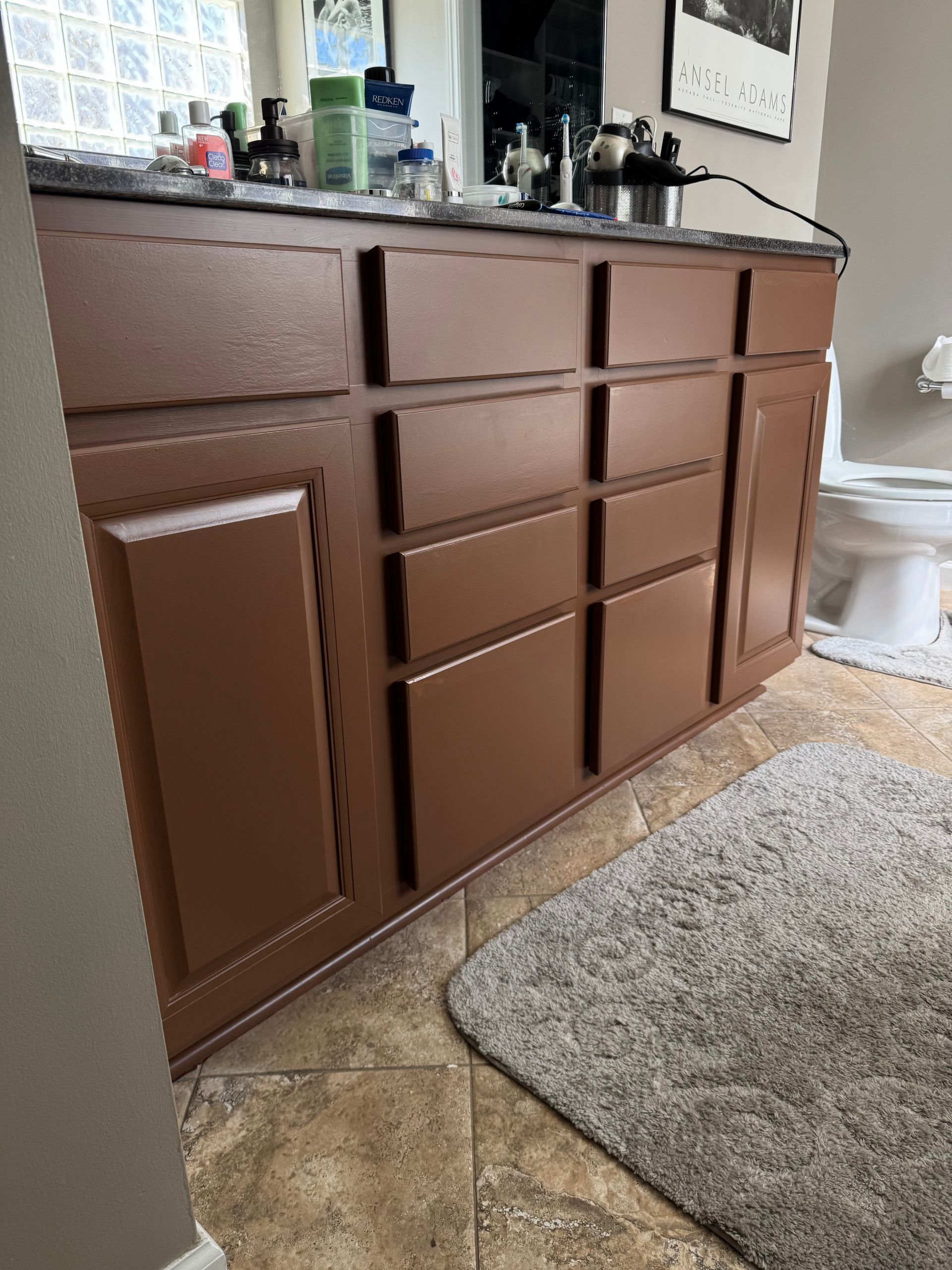 Brown bathroom vanity with drawers and doors, countertop with toiletries, and a gray rug on the floor.