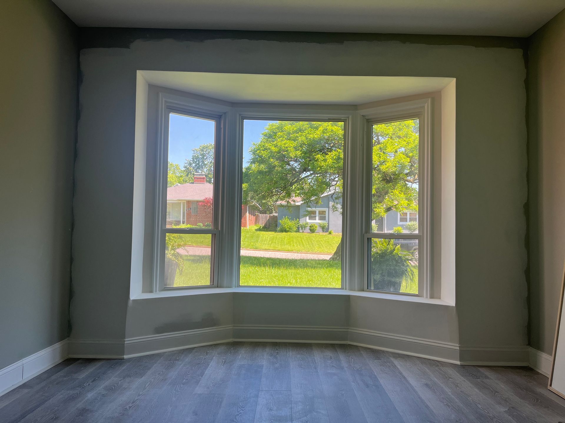 A bay window with three panes, looking out onto a sunny yard. Room with gray walls and wood flooring.