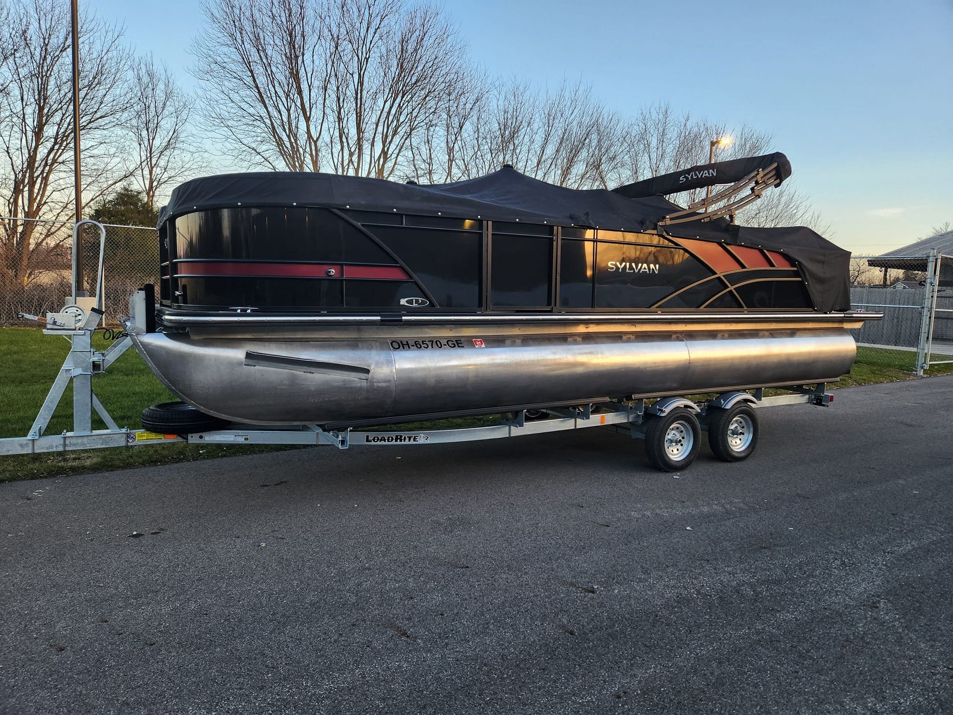 A black and bronze pontoon boat with a cover, sitting on a boat trailer on asphalt.