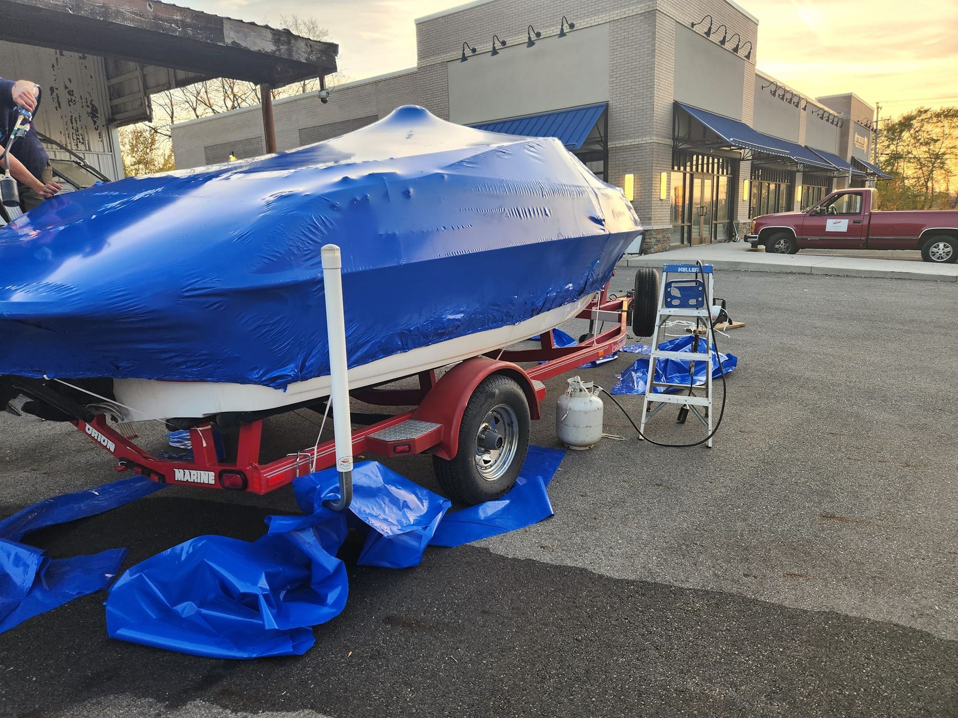 A boat covered in blue shrink wrap sits on a red trailer in a parking lot, with a ladder and propane tank nearby.