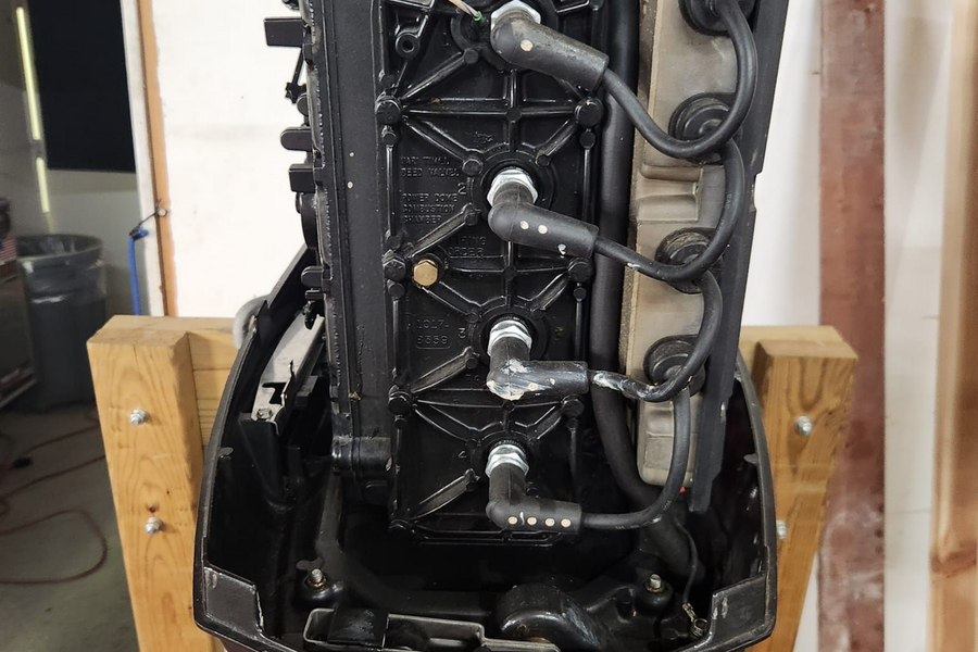 A black marine outboard motor engine mounted on a wooden stand, showing four spark plug wires connected to the block.
